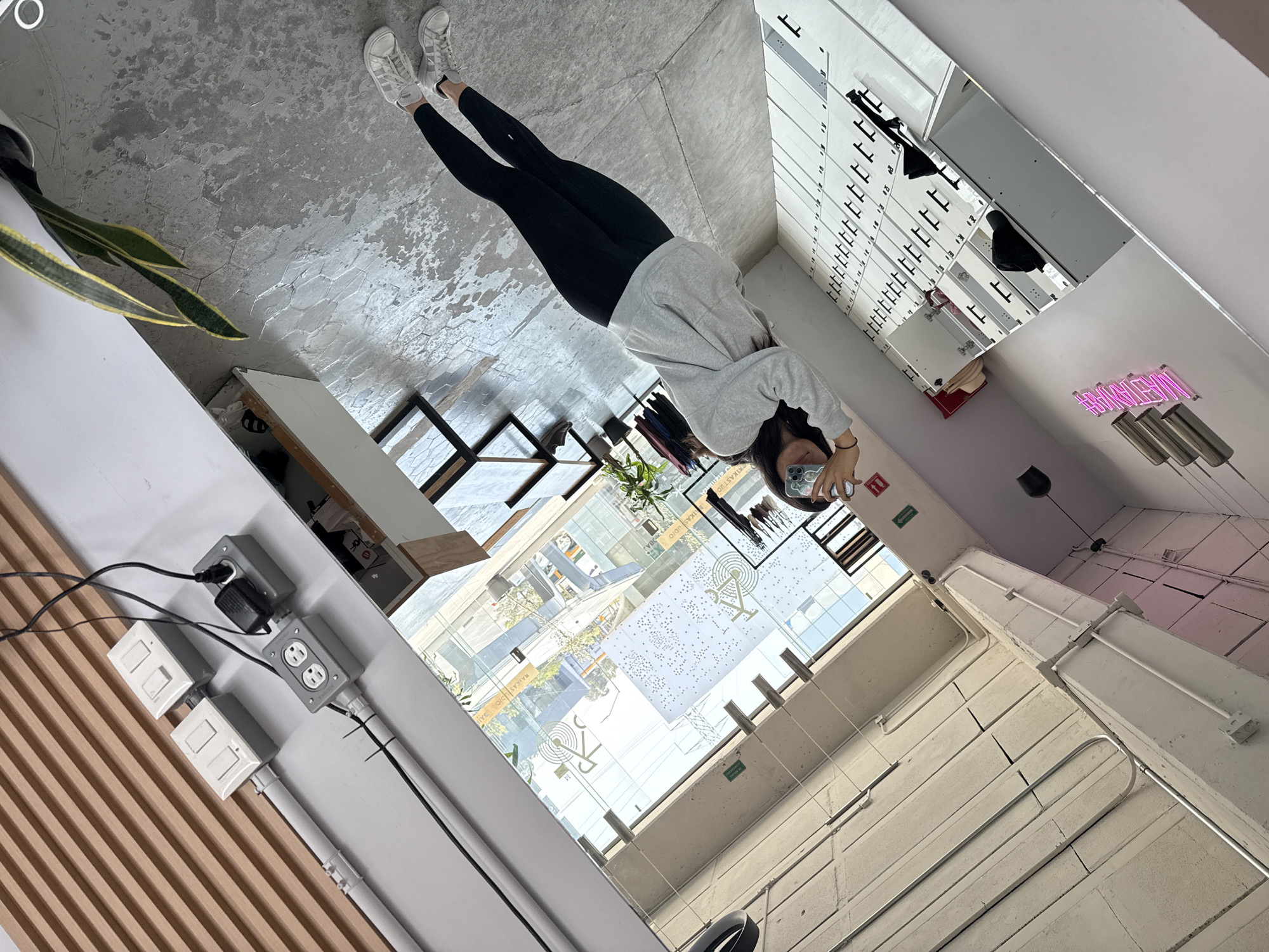 The image captures a person taking a mirror selfie in a modern, industrial-style indoor setting, likely in an urban area during daytime. The person, positioned centrally, is dressed in white Adidas sneakers, black leggings, and a light grey hooded sweatshirt. They are holding a blue/silver smartphone, partially obscuring their face, to capture their reflection in a large, wall-mounted mirror.
The floor is a textured grey surface with a subtle hexagonal pattern, resembling concrete. To the left, a plant with long green and yellow leaves is visible in a pot, near a white counter or table. On the right, the prominent mirror reflects various features of the room. This includes a series of white storage units or lockers with black handles and numbered labels (such as '1', '11', '12', '13'), along with a bright pink neon sign displaying "HAGYM" (reversed in the reflection). Metallic pendant lights are suspended below the neon sign.
Through large windows and possibly skylights, a clear, sunny outdoor scene is visible, showing distant buildings, trees, and what appears to be a city street or courtyard. Some window panels feature decorative white and gold decals, including reversed text that reads "RAKAUDIO". The walls and ceiling are light-colored, constructed from concrete blocks, with exposed electrical conduits running along them. Near the bottom left, a section of light brown slatted wooden paneling can be seen, adjacent to grey electrical boxes containing outlets (one with a charger plugged in) and white light switches. A red and white information or exit sign (reversed) is also faintly visible in the reflection. The overall environment suggests a contemporary gym, studio, or commercial space.