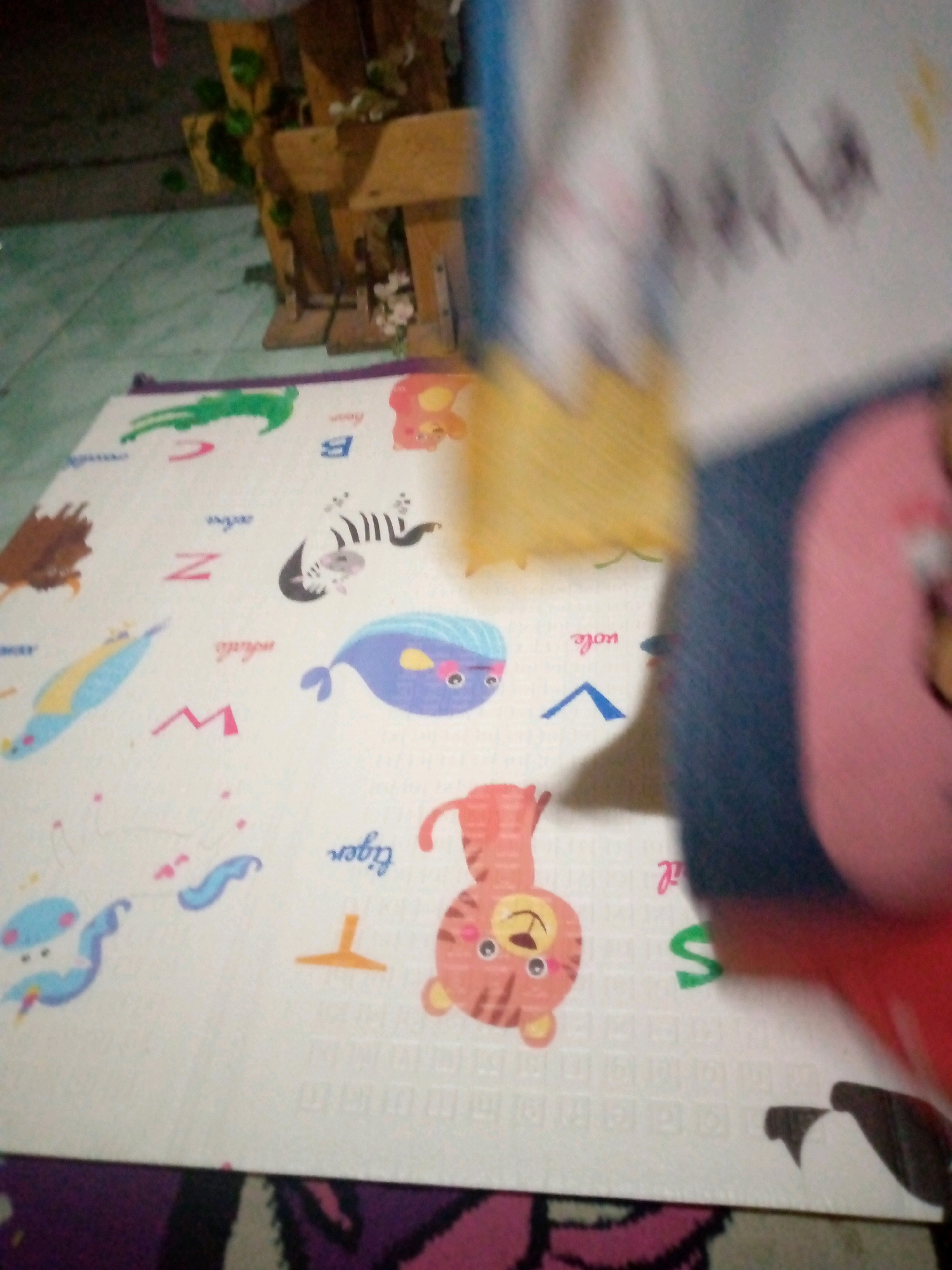 The image displays a children's playmat with alphabet letters and cartoon animal illustrations. The playmat is predominantly white with colorful designs. To the left of the mat, a green alligator is depicted next to the letter 'C'. Further down, a brown, furry creature resembling a hedgehog or a bird is shown. In the center, a zebra is illustrated next to the letter 'Z'. Below the zebra, a blue whale with orange details is visible. Towards the bottom right, a tiger character is featured, and to its left, the word "tiger" is written in cursive. A blue, unicorn-like creature with a yellow horn is also present on the left side. The playmat is laid out on a green tiled floor. In the background, a wooden shelf structure with some greenery and white flowers is visible. The overall scene suggests an indoor setting, likely a home or play area. The location is identified as Sumberwono, Indonesia. The image is blurry and partially obscured by a dark, out-of-focus object in the foreground, possibly a toy or a person.