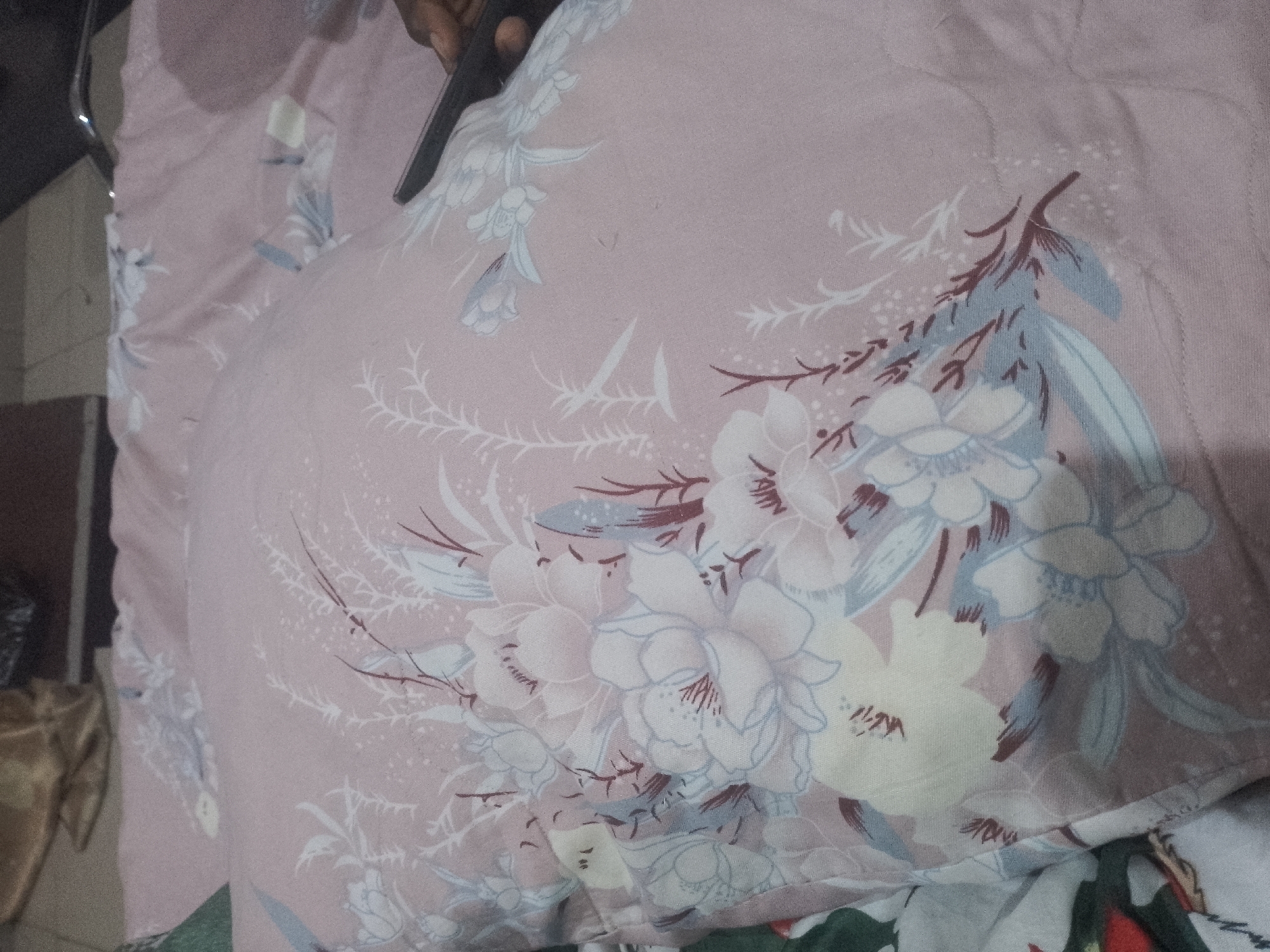 A close-up, slightly angled shot shows a person's hand holding a black object over a pink floral patterned fabric. The fabric appears to be a pillow or duvet cover, with large white and blue flowers and delicate branches with brown accents. The lighting is dim, casting shadows and highlighting the texture of the fabric. The background is out of focus, with hints of other materials and possibly furniture. The scene is indoors.