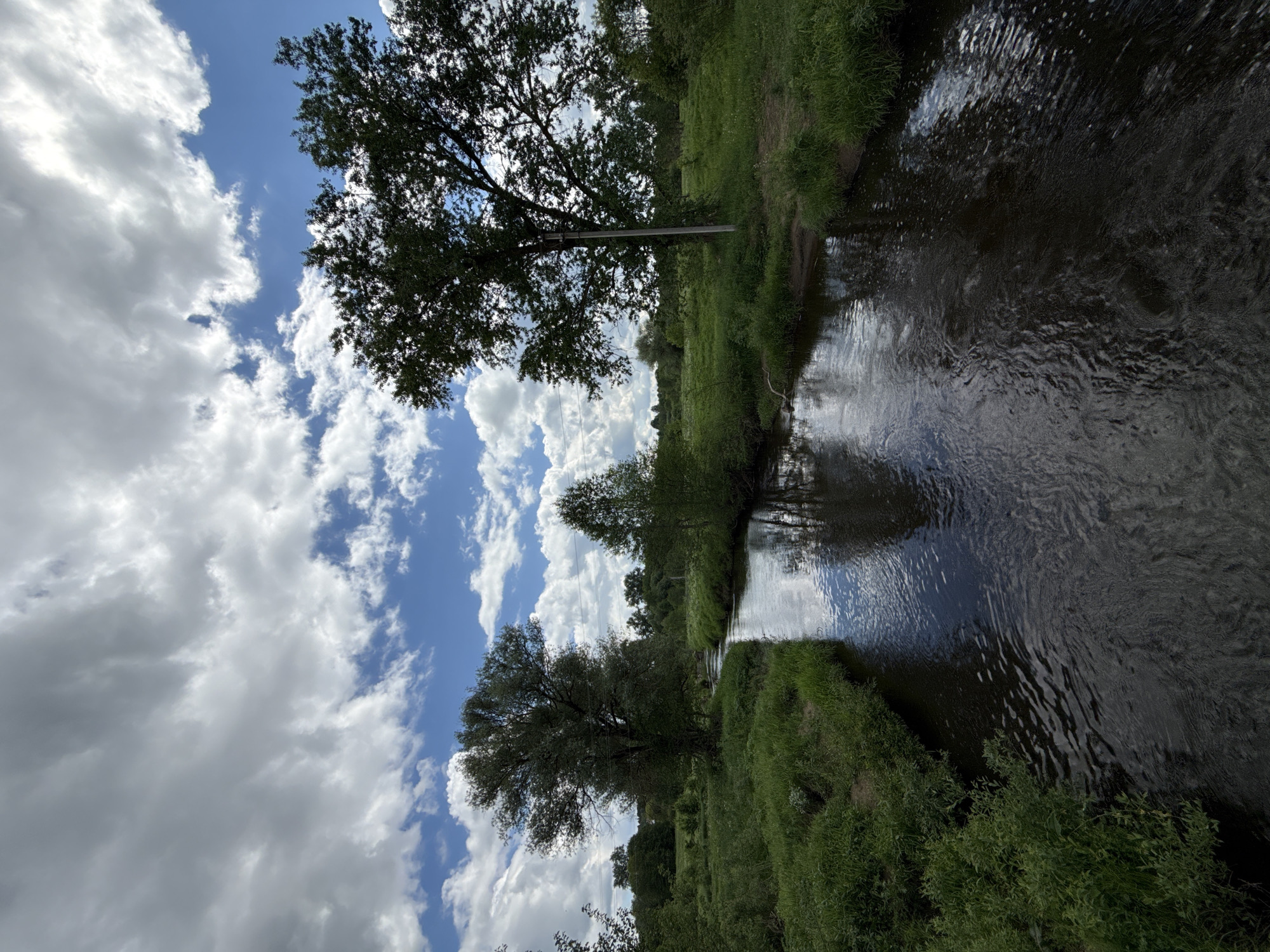 The image depicts a narrow river flowing through a green landscape under a cloudy blue sky. The location is Уральское, Russia. Trees line the banks of the river, and their reflections are visible in the water. Power lines run through the trees. The vegetation along the river is lush and green.