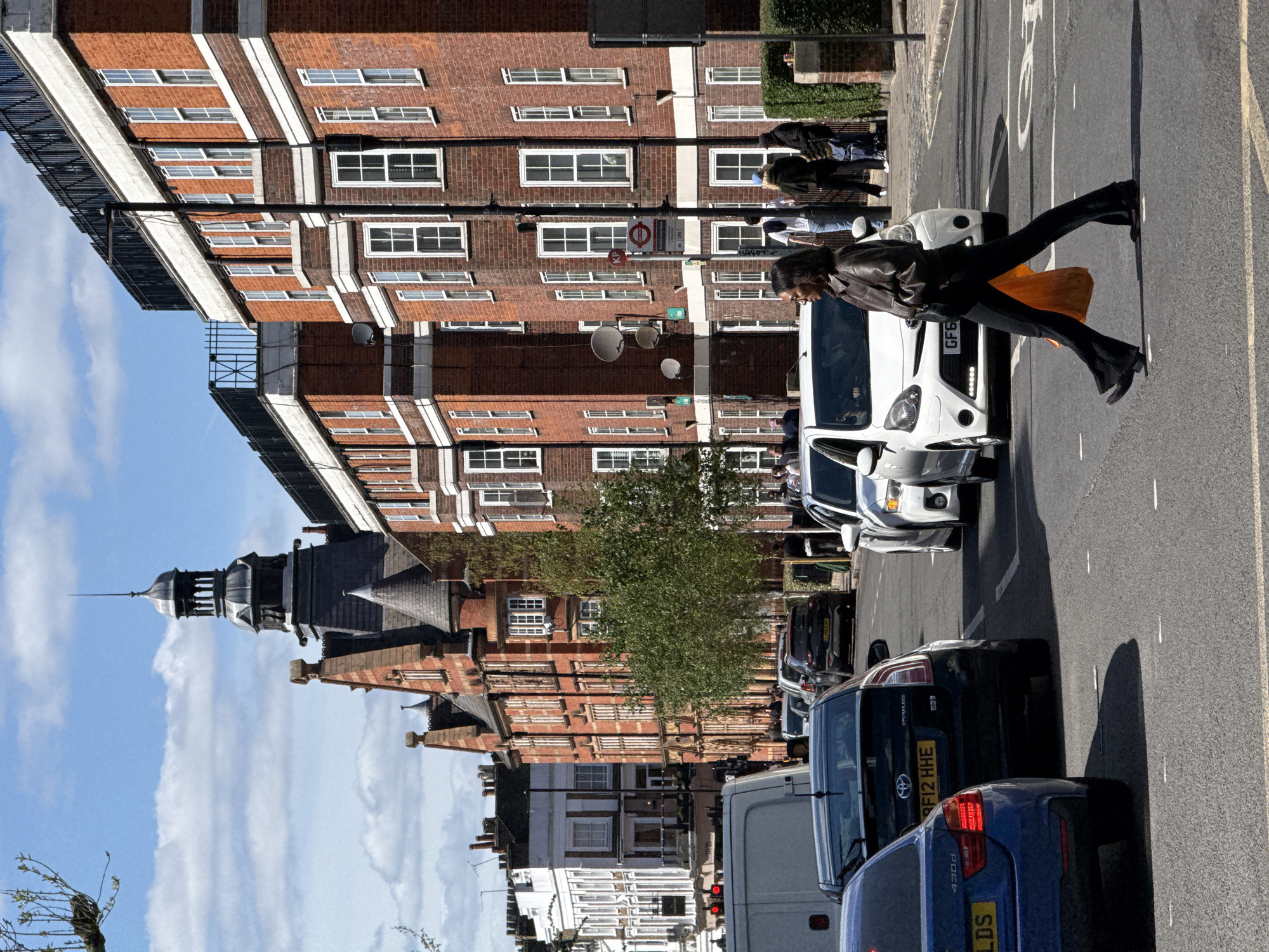 A street scene in London, United Kingdom, oriented vertically, shows a woman crossing the road in front of a white van. The woman wears a brown leather jacket, black pants, and black boots, and carries an orange bag. Several cars are parked along the side of the road, including a black car with the license plate "FZ12 HHE" and a blue car. Buildings line the street, consisting of brick apartment buildings with white-framed windows, interspersed with a tower with a dark roof and spire. A London Underground sign is visible mounted to one of the buildings. Pedestrians can be seen walking on the sidewalk near the apartment buildings. The sky is blue with scattered clouds.