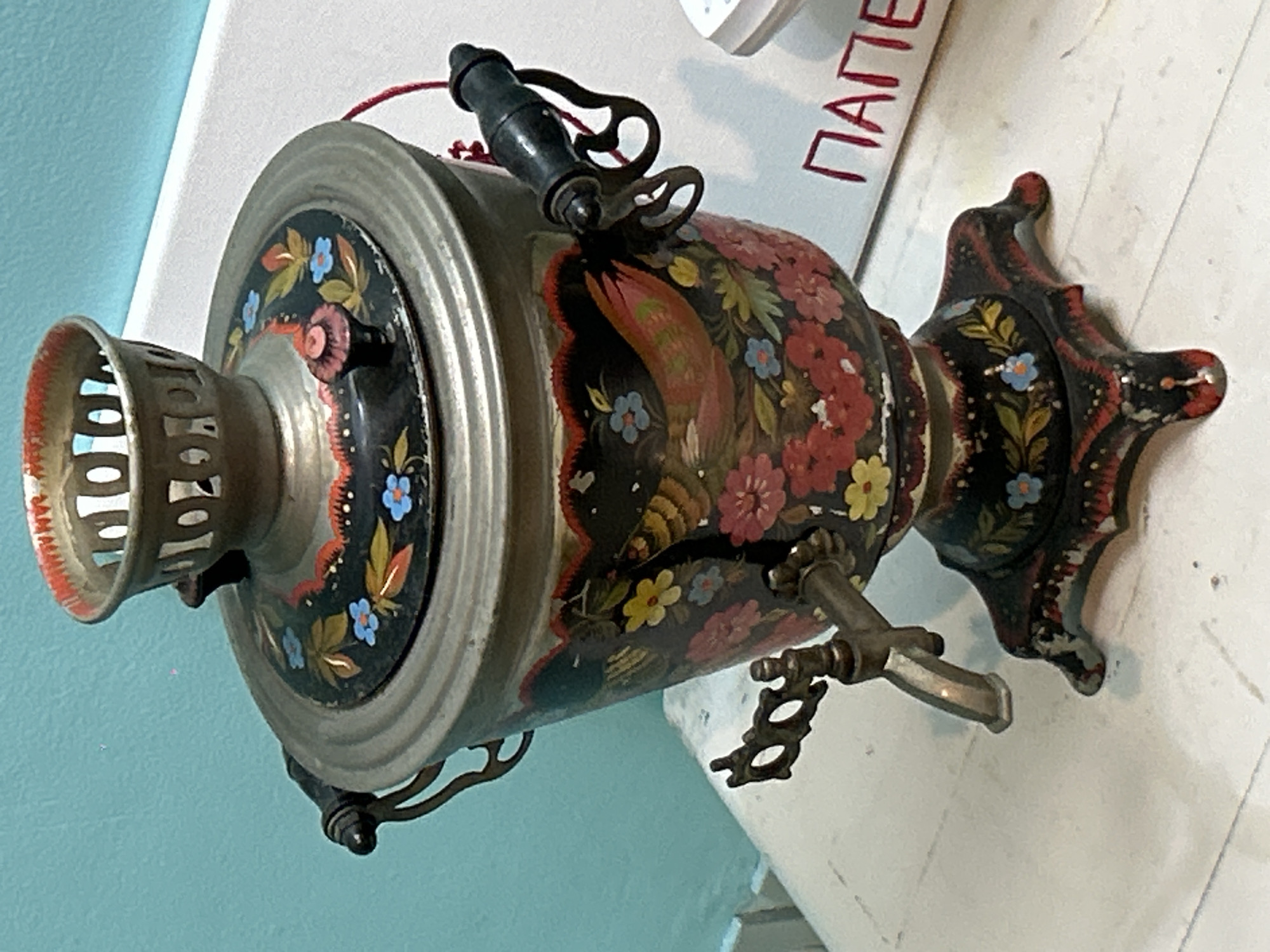 The image shows a vintage samovar, elaborately decorated with floral and bird motifs in vibrant colors against a dark background. The samovar features a metallic finish, a spigot with an ornate handle, and a decorative top with a black handle. It appears to be resting on a white surface near a wall painted in light blue. There is also a magazine or pamphlet with a red inscription on it. The location in North Kuta, Indonesia, is not verifiable from the image alone.