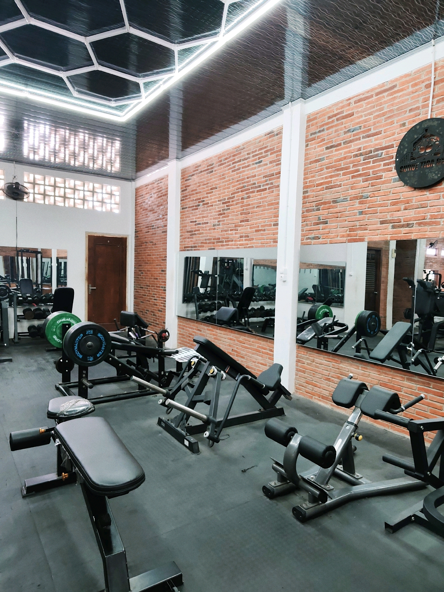 The image depicts the interior of a gym, likely located in Pancur, Indonesia. The gym features a modern design with exposed brick walls and a hexagonal ceiling with integrated lighting. Various weightlifting equipment is visible, including weight benches, barbells with weight plates, and what appears to be a leg press machine. Mirrors line the walls, reflecting the gym equipment and adding to the sense of space. The floor is covered in black mats, typical for a gym environment. There are no people visible in the image. The overall impression is that of a well-equipped and clean fitness facility.