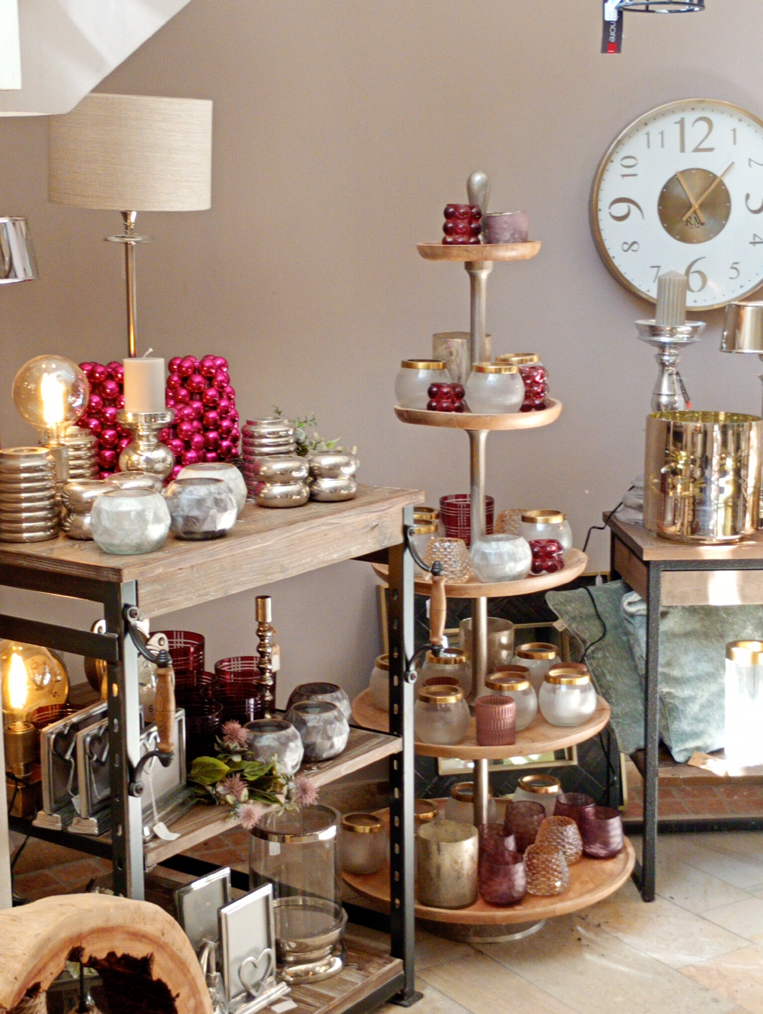 The image depicts an interior scene, possibly a retail store, showcasing various decorative home items. The room has light brown walls and a light-colored ceiling with a sloped section, suggesting an attic or an angled architectural feature. The floor is made of light-colored wood planks.
In the foreground, a rustic wooden table is laden with silver and glass decorative objects, including candle holders, vases, and what appear to be small decorative balls in a vibrant pink color. To the left of this table, a table lamp with a textured beige shade is visible.
Behind the table, a large white wall clock with gold numerals is mounted on the wall. To the right of the clock, a tiered display stand holds more decorative items, including glass jars, candle holders, and what seem to be small red decorative spheres. Further to the right, a tall, metallic gold cylindrical object, possibly a vase or a large candle holder, is placed on a dark surface.
Several shelves are present, holding a variety of glass and metallic decorative pieces. One shelf on the left displays framed pictures, one with a heart design. Another shelf lower down has red glass containers and metallic candle holders. The overall impression is that of a well-arranged display of home decor items.
The location is identified as Bad Sachsa, Germany, and the scene is likely within a commercial establishment. There are no people visible in the image. The lighting suggests an indoor setting with artificial and possibly natural light contributing to the ambiance. The items are arranged in a manner that emphasizes their decorative qualities.