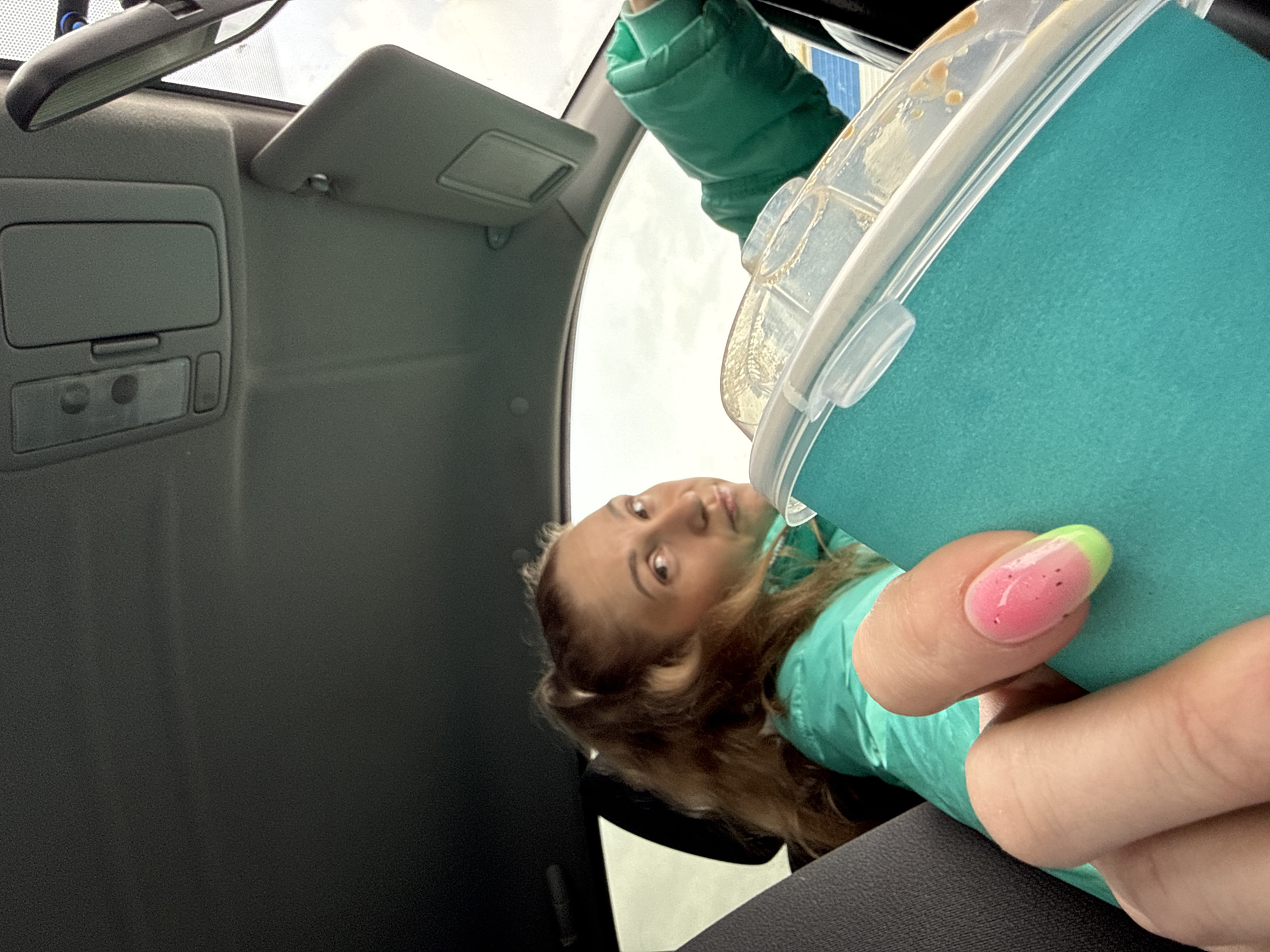 A photograph taken from the interior of a vehicle, possibly a car, depicts a young woman holding a turquoise cup with a clear lid. The woman, with light brown hair, is wearing a light green jacket. Her right thumb is visible, featuring a manicure with pink and green accents. The interior of the vehicle is gray, with the sun visor open. The scene suggests the vehicle is parked, possibly in Chaikovsky, Russia, though this cannot be confirmed visually.