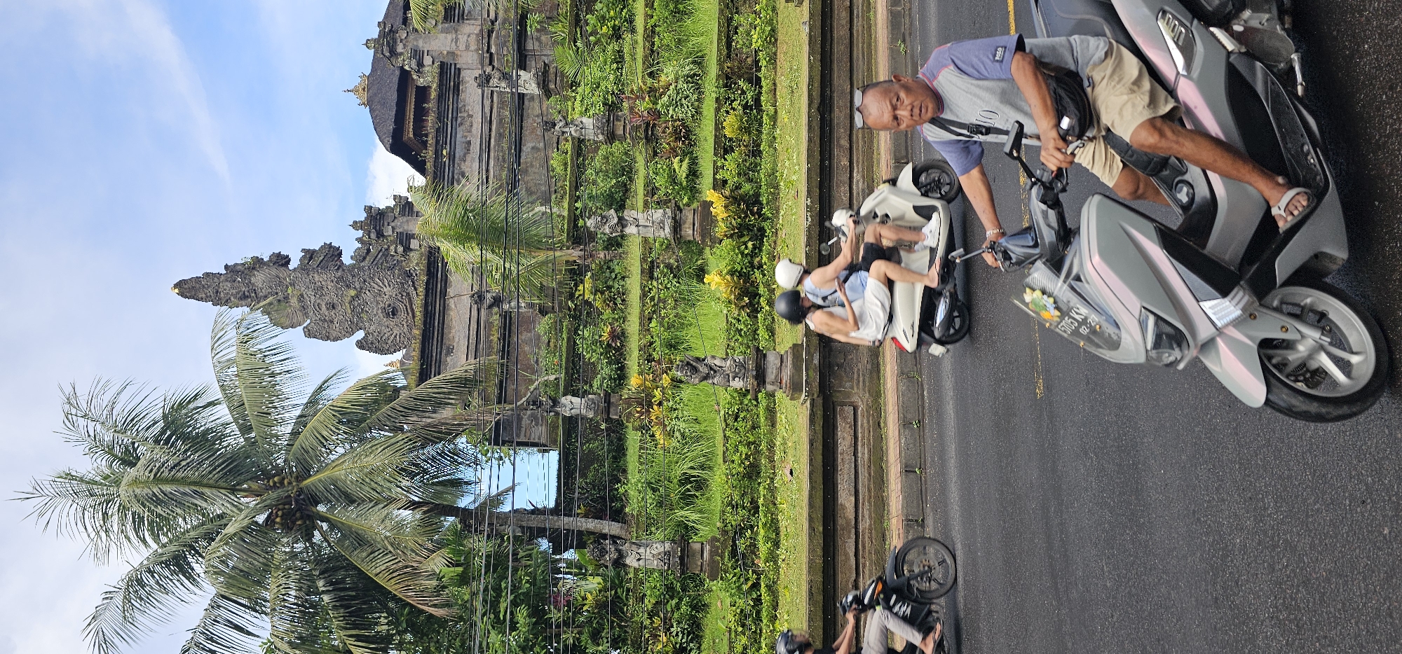 The image shows a scene in the Ubud District of Indonesia. A traditional Balinese temple structure is partially visible amidst lush greenery and a coconut tree. On the adjacent road, there are people on mopeds. One moped has two people riding tandem while a third person stabilizes the vehicle. A second moped is further down the road.
