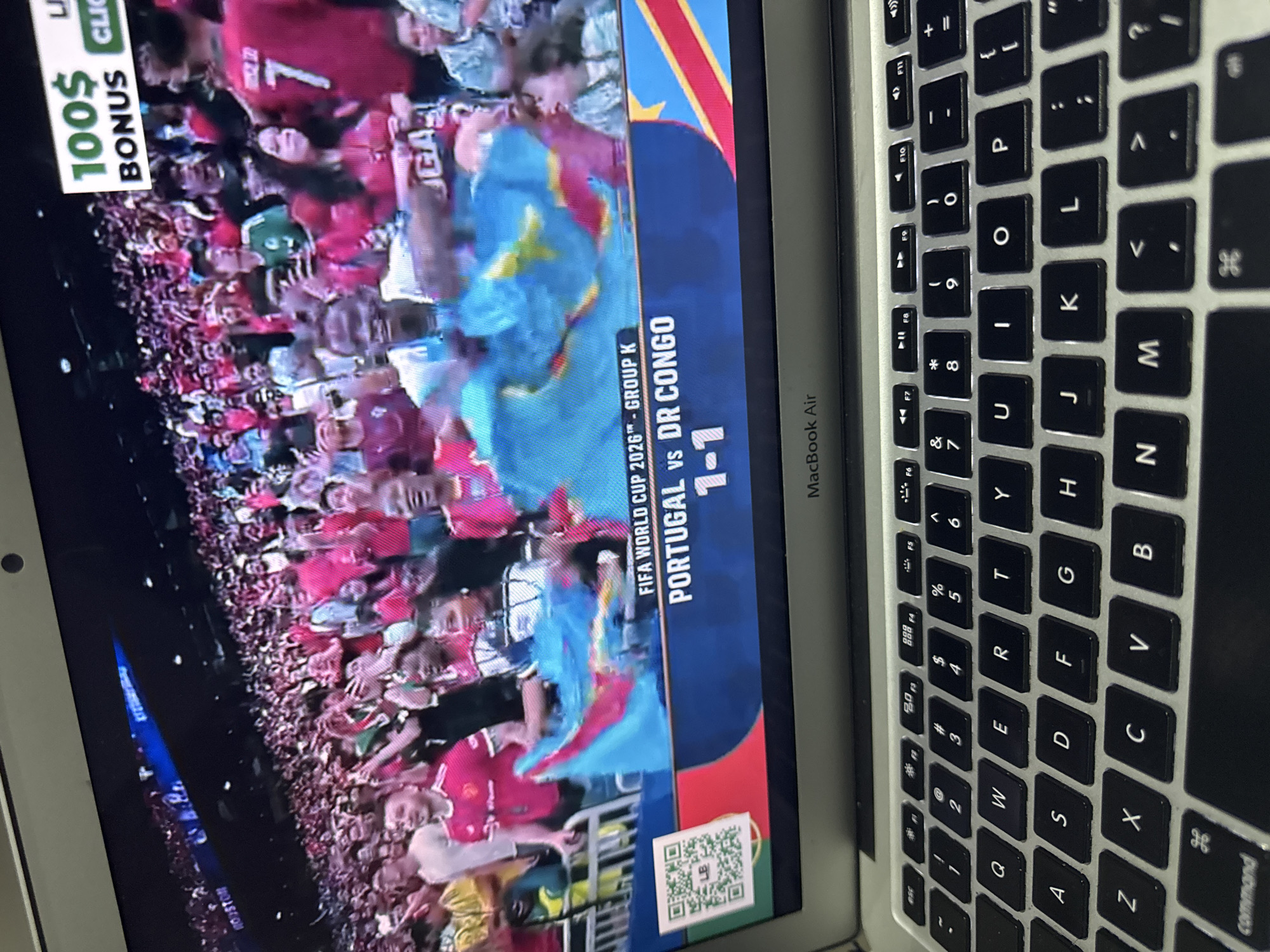 The image shows a laptop displaying a soccer match broadcast. The screen shows a live score graphic for "FIFA WORLD CUP 2026™ - GROUP K" with "PORTUGAL vs DR CONGO" and the score "1-1". A crowd of spectators is visible in the background, many wearing red or blue jerseys. There is also an advertisement banner at the top of the screen that reads "100$ BONUS". The laptop itself is open and on a surface. The location of Kufena, Nigeria is not depicted or referenced in the image.