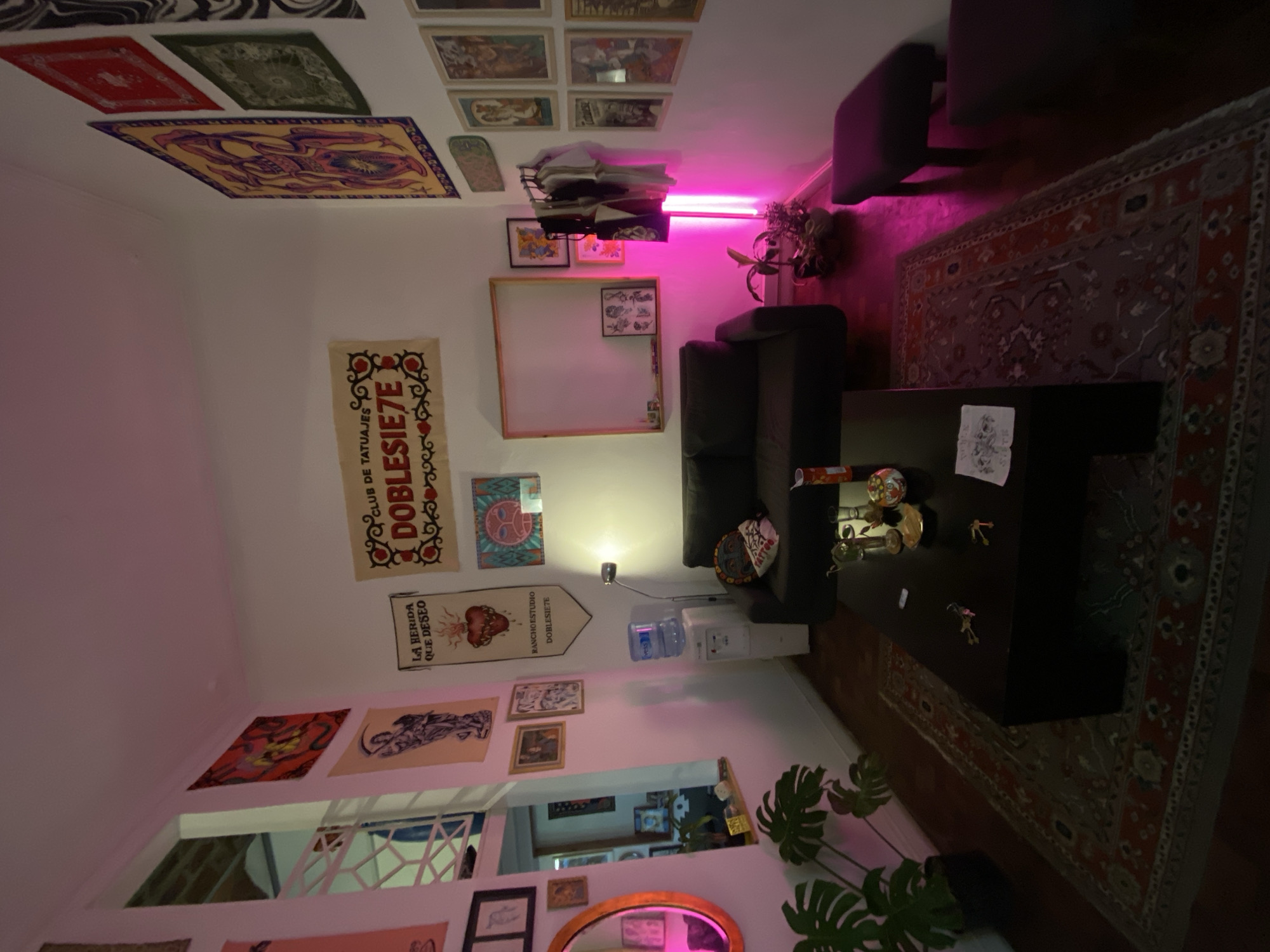 The image depicts an interior space, likely a tattoo studio or artistic workspace, located in Buenos Aires, Argentina. The room is decorated with various tapestries, posters, and artworks displayed on the walls. A white board with a wooden frame is visible near a black sofa. A coffee table sits in front of the sofa, cluttered with objects including decorative skulls and keys. A water dispenser is placed against the wall, near the sofa. The floor is covered by a red and grey ornate rug. Light from an unseen source casts a pink hue throughout the scene. A houseplant is located near a mirror. The presence of the banners with Spanish text suggests this is somewhere in the city of Buenos Aires, Argentina, where Spanish is the primary language.