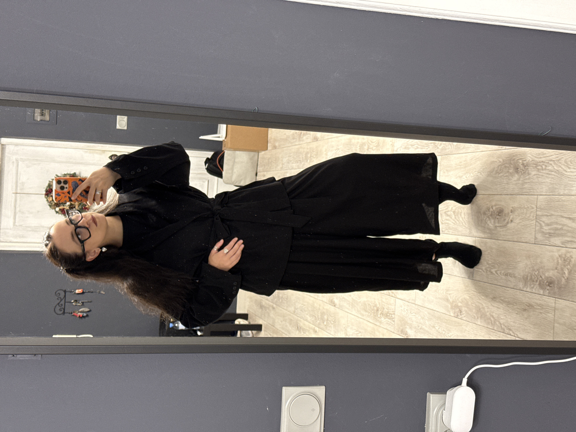 A full-body, mirrored selfie shows a woman wearing black clothing. She has dark, wavy hair pulled back into two ponytails and is wearing black-rimmed glasses and heart-shaped earrings. Her outfit consists of a belted blazer over a dark top and wide-leg pants. She has on black socks. The woman is standing in what appears to be a domestic setting, possibly an apartment. In the background, a door with a wreath is visible, and to the left, a white electrical outlet with a charging cable is mounted on a gray wall. The floor is light-colored wood. The image does not provide any information that would suggest a location in Chaikovsky, Russia.