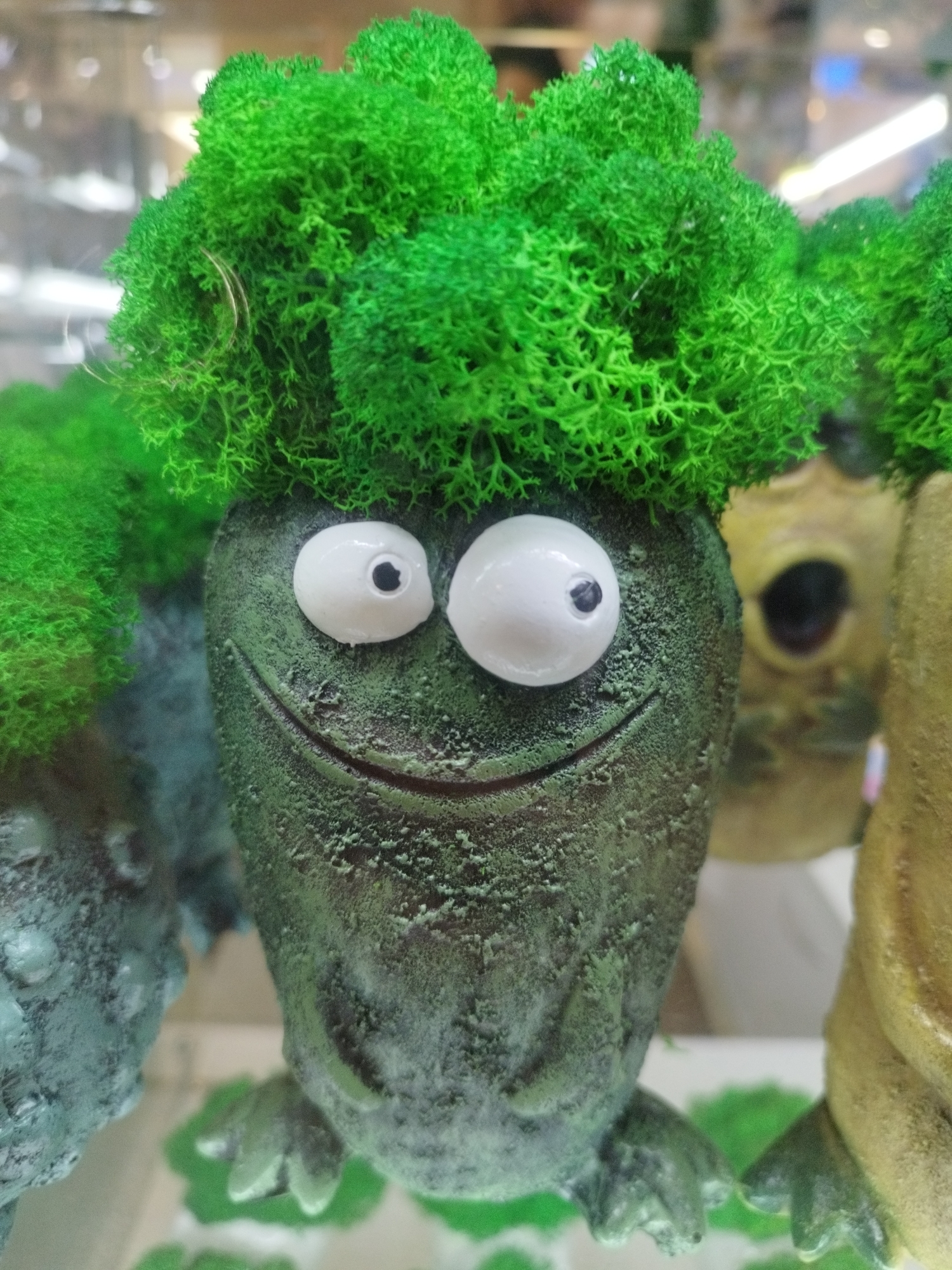 This is a close-up, eye-level, medium shot of a decorative object resembling a frog with moss for hair. The object is green with a rough, textured surface. It has two large, white, cartoonish eyes with black pupils. A wide, carved smile is visible on its face. The object is adorned with bright green, fluffy moss that resembles hair and is also present on its feet and as surrounding decorative elements. In the blurred background, other similar decorative objects are visible, one appearing yellow and another with a bluish-green hue. The scene is indoors, likely in a shop or display setting, given the reflections of lights and shelves in the background. No people are present, and no specific location in Saint Petersburg, Russia, can be identified from the image.