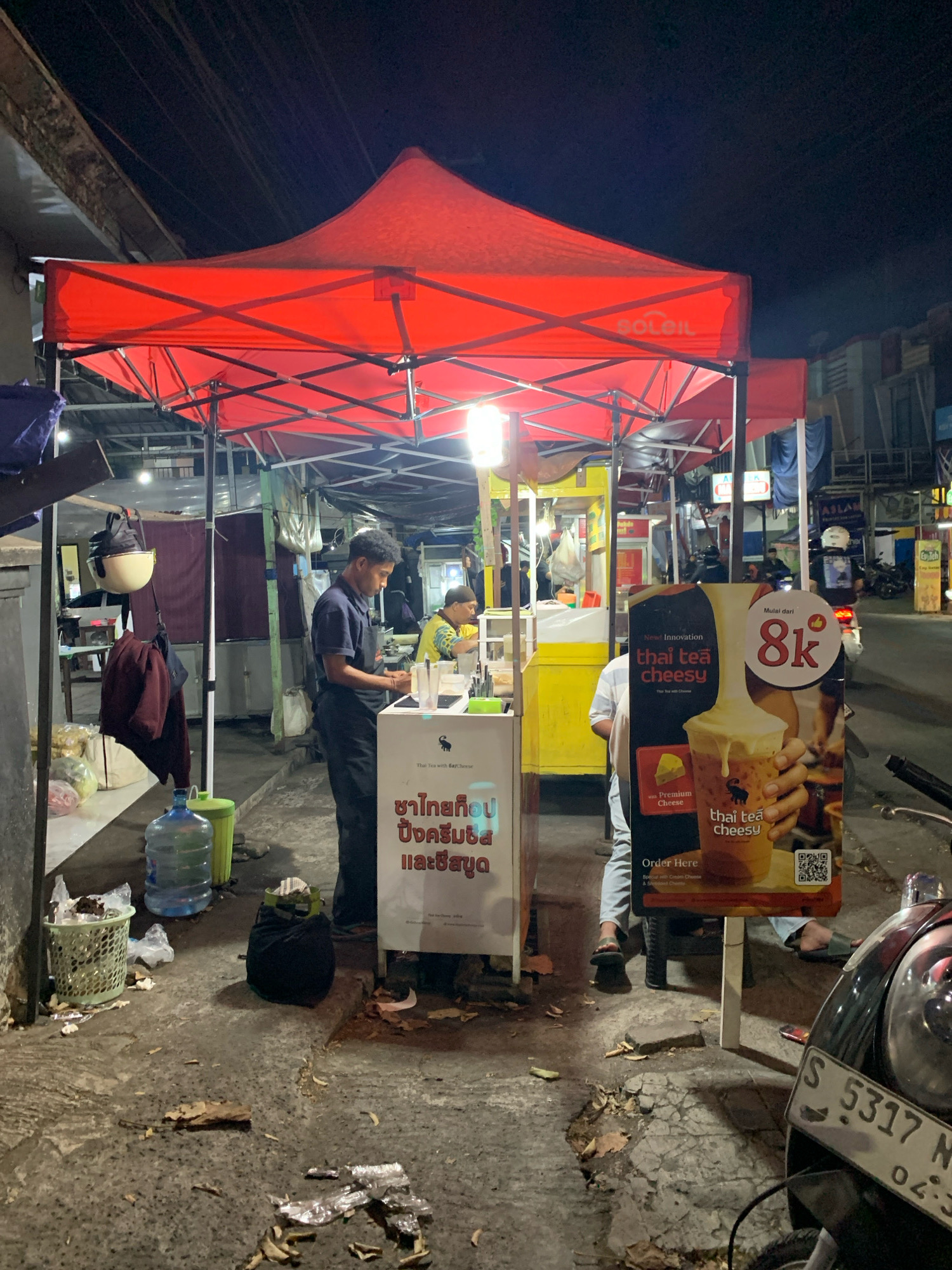 An outdoor food stall operates at night in Malang, Indonesia. A red canopy tent shelters the stall, which appears to be selling Thai tea. A man in a dark blue shirt and black pants is preparing drinks behind a counter. To his right, a large sign advertises "Thai Tea Cheesy" with an image of a drink and a price of 8k. Another smaller sign in front of the stall has text in what appears to be Thai script. A younger person in a yellow shirt is seated further back in the stall, looking down at something on the counter. In the foreground, a motorcycle is parked, and the ground is littered with debris, including leaves and food wrappers. Several large water bottles and a trash can are also visible near the stall. The background shows a street scene with other stalls and buildings illuminated by artificial lights.