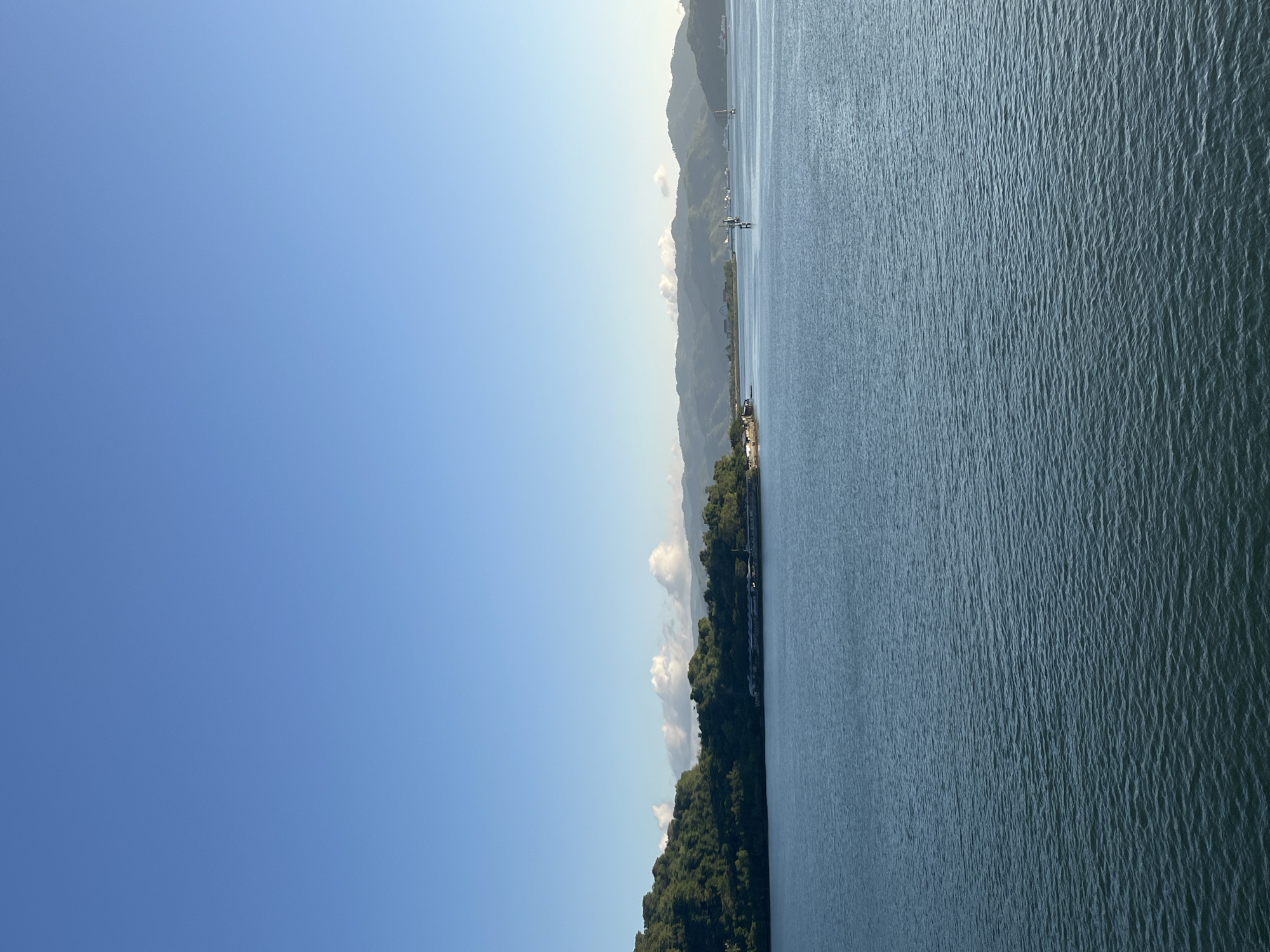A panoramic view of a body of water with a lush, tree-covered island in the distance, Batas Desa Skotong Timur, Indonesia. The sky is a clear, bright blue. The water is calm with small ripples. On the island, there is a long, low-rise building that appears to be a pier or a marina. Further in the distance, mountains are visible. There are a few small boats or buoys scattered on the water. The overall scene depicts a serene coastal landscape under a clear sky.