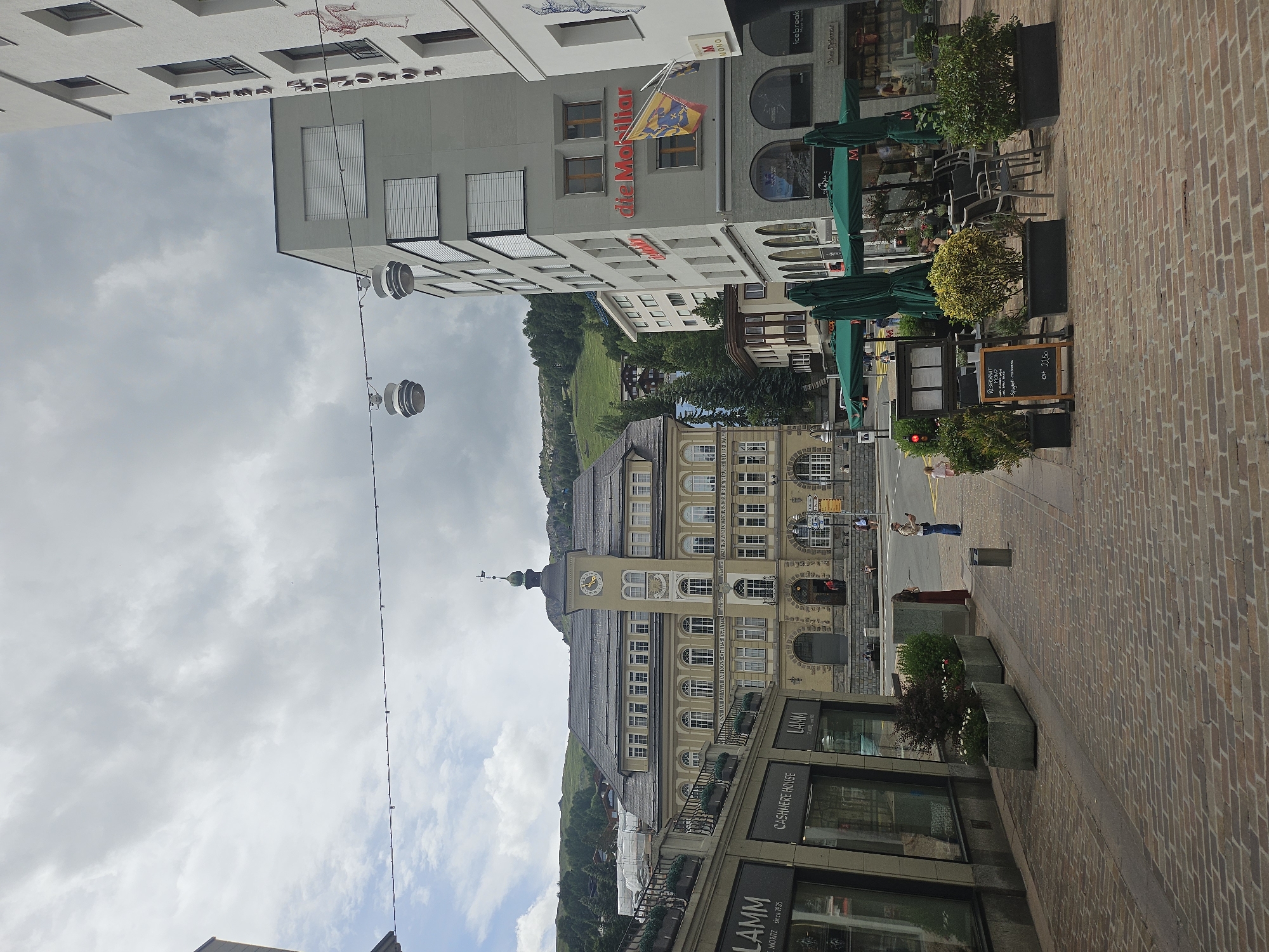 This is a slightly rotated, medium shot of a street scene in St. Moritz, Switzerland. The scene is dominated by buildings on the left and a cobblestone street on the right. Several people are visible walking on the sidewalk and street.
The buildings on the left appear to be a mix of modern and older architecture. One building has the text "die Mobiliar" and "M MONO" visible, along with a flag. Another building shows "CASHMERE HOUSE" and "LAMM" in large letters. A sign for "RESTAURANT" and "Spaghetti carbonara CHF 22.50" is also visible. The architecture suggests a European town, possibly in the Alps, with a clock tower visible on one of the buildings. The sky is overcast, suggesting cloudy weather.
The street is paved with tan cobblestones and lined with planters containing green shrubs and some flowers. Green umbrellas are set up outside what appears to be a cafe or restaurant. The overall impression is of a bustling street in a picturesque European town, likely during the daytime, given the lighting.