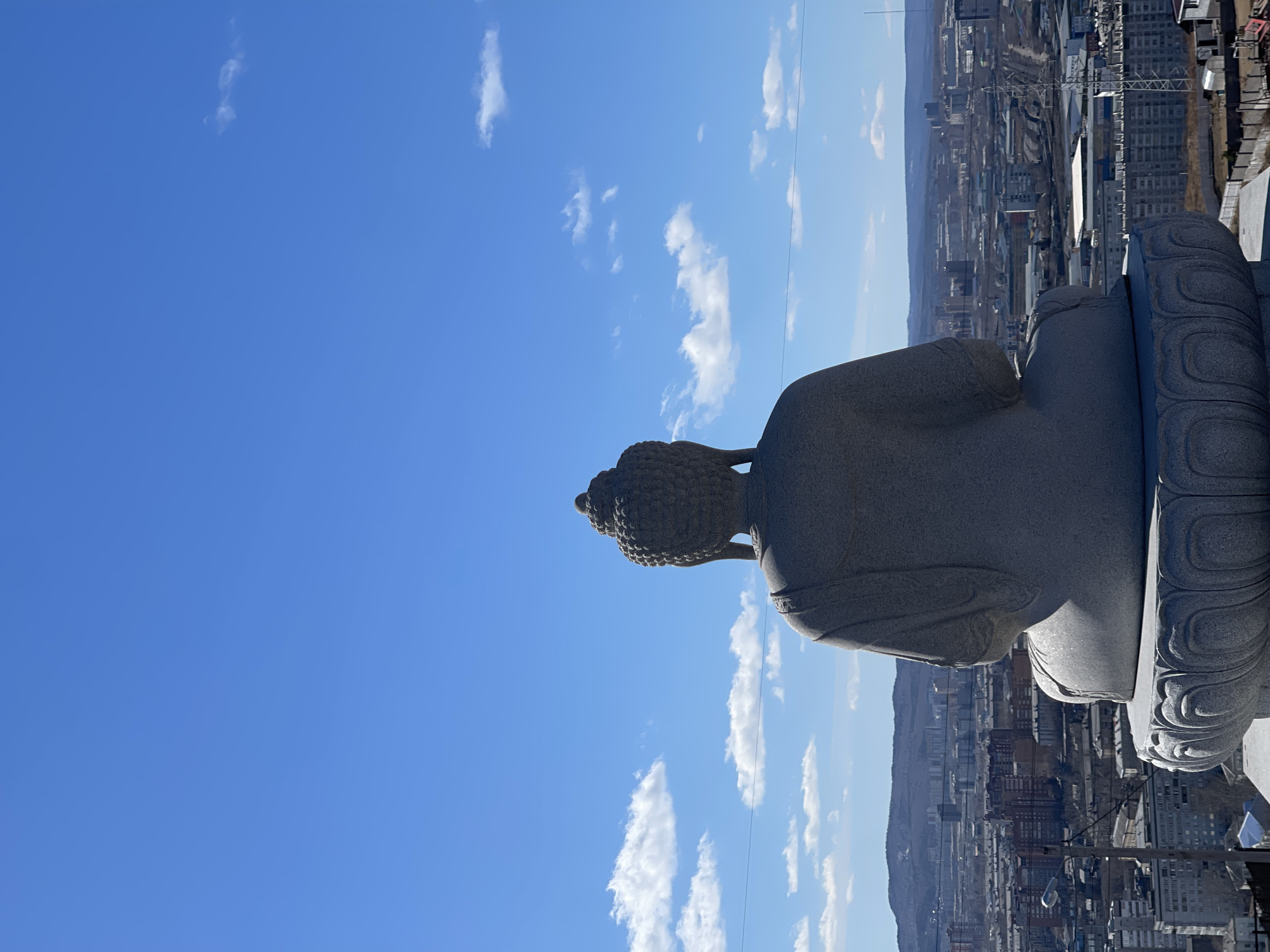 A statue of Buddha sits on a platform overlooking the city of Krasnoyarsk, Russia. The statue is stone, with a textured head and smooth body. It sits on a decorated base. The background shows a sprawling urban landscape with a mix of residential and industrial buildings under a blue sky dotted with white clouds.