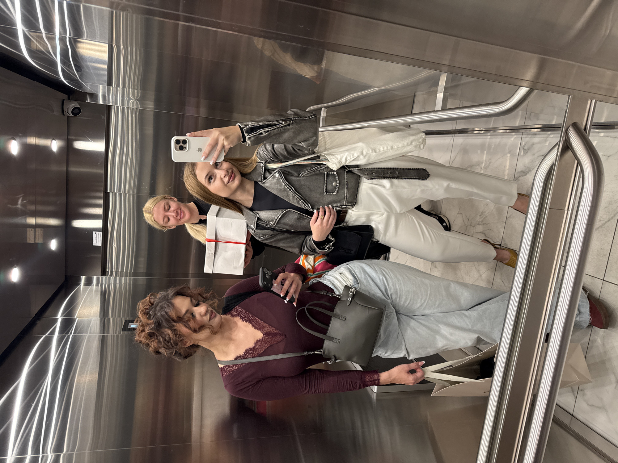 Three women are in an elevator taking a selfie. The woman closest to the mirror is holding a smartphone, likely to capture the group selfie. She has long blonde hair, wears a black top under a distressed denim jacket, white pants, and is accessorized with a black purse. Her friend in the background has blonde hair in a ponytail and is holding a white gift box with a red ribbon. In the foreground, the third woman, with curly brown hair, is in a long-sleeved burgundy lace-trimmed top and light blue jeans. She is holding a smartphone and has a gray handbag. They are standing on a marble floor in a metallic elevator. The presence of safety rails in the elevator is visible. The location cannot be determined to be in Belgrade, Serbia based on the image alone.