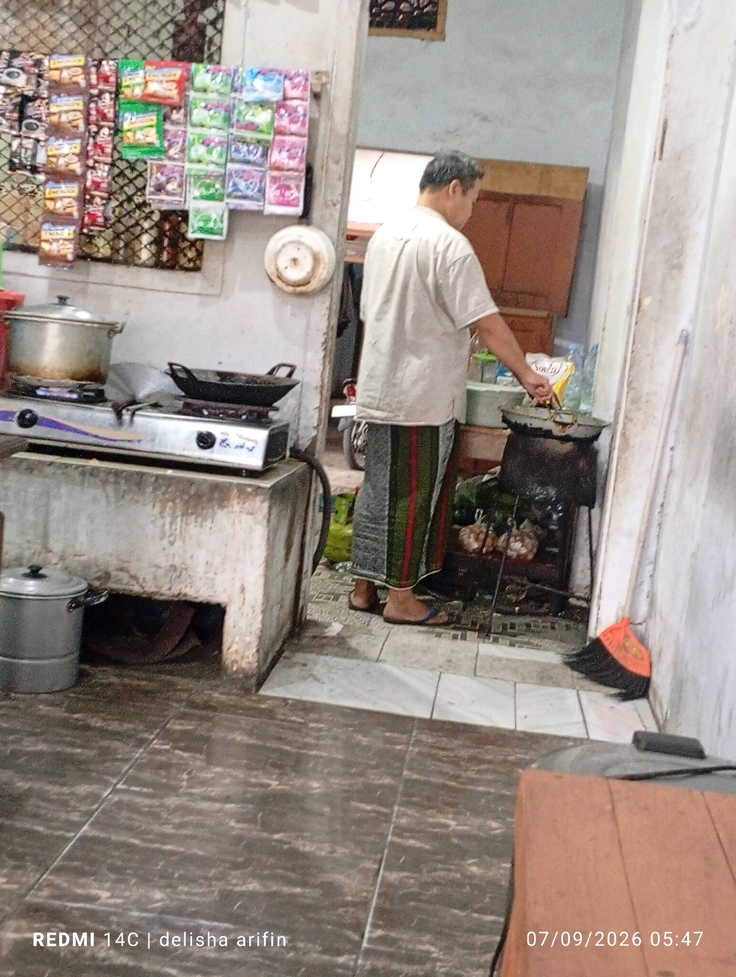 In a dimly lit, enclosed outdoor space, likely a kitchen or food stall area in Gambir Kuning, Indonesia, a man is cooking over an open flame. He is wearing a light-colored short-sleeved shirt and a sarong with green and red stripes. He is standing and holding a wok or frying pan with a utensil, seemingly stirring its contents.
The environment is utilitarian and appears to be a working kitchen. To the man's left, a stainless steel gas stove with two burners is visible, with a large pot on the left burner and a black frying pan on the right. This stove is placed on a rough, concrete counter. Below the counter, a metal pot with a lid is situated on the ground.
Behind the man, a small shelf or stand holds several items, including what appear to be bags of food products hanging on the wall above. Further back, there's a glimpse of what looks like a motorcycle and green gas cylinders. To the far right, an orange and black broom is leaning against a wall. The floor is tiled with a dark, patterned material. The overall impression is one of a busy, functional cooking area.