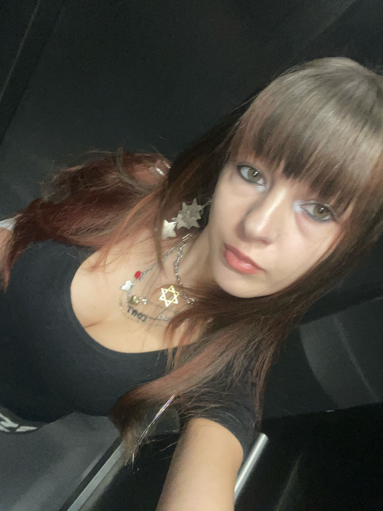 The image is a close-up, selfie-style photograph of a young woman with long, reddish-brown hair and bangs. She is wearing a black, low-cut t-shirt and several necklaces, including one with a Star of David pendant. Her makeup includes prominent eyeliner and lip gloss. The background is dark and out of focus, suggesting an indoor environment, possibly a car or a dimly lit room. There are no specific landmarks or elements that would indicate a location in Raanana, Israel.