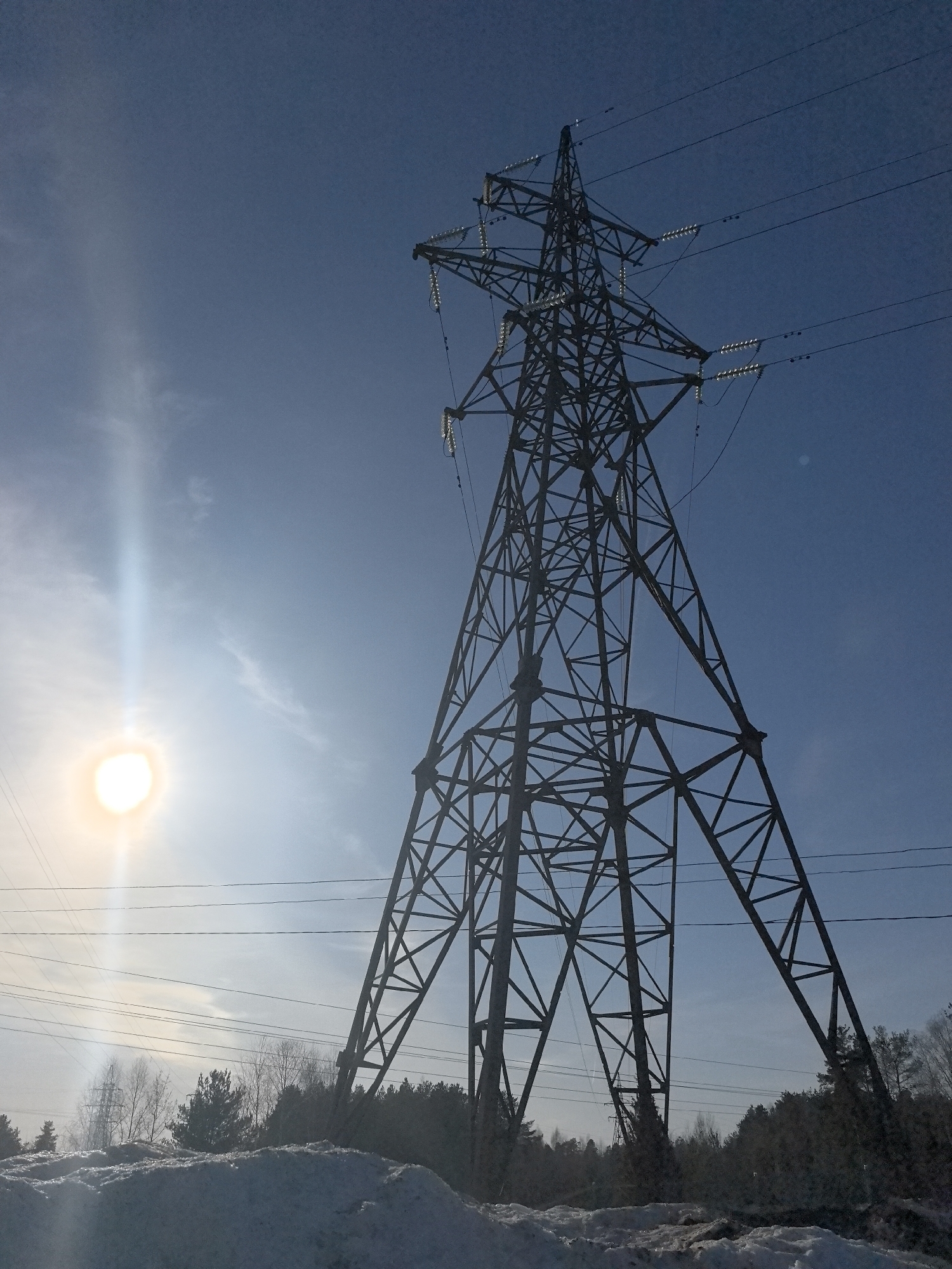A high-angle, wide shot shows a tall, dark metal electricity pylon standing against a blue sky with a bright sun to the left. The pylon is an intricate framework of metal beams, supporting insulators and power lines. In the foreground, the ground is covered in snow. Trees and other vegetation are visible behind the snow-covered ground. The sun's glare creates a vertical streak in the sky. This image could be taken at Noviy, Russia given the presence of snow and power lines.