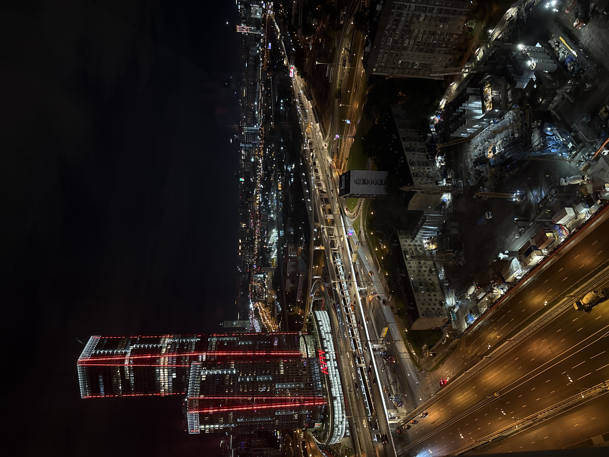 The image depicts a nighttime view of Khimki, Russia, from an elevated perspective. The scene features multiple illuminated highways with car lights visible, a partially completed construction site with visible equipment and cranes, and a high-rise building with red lighting accents. City lights stretch into the distance.