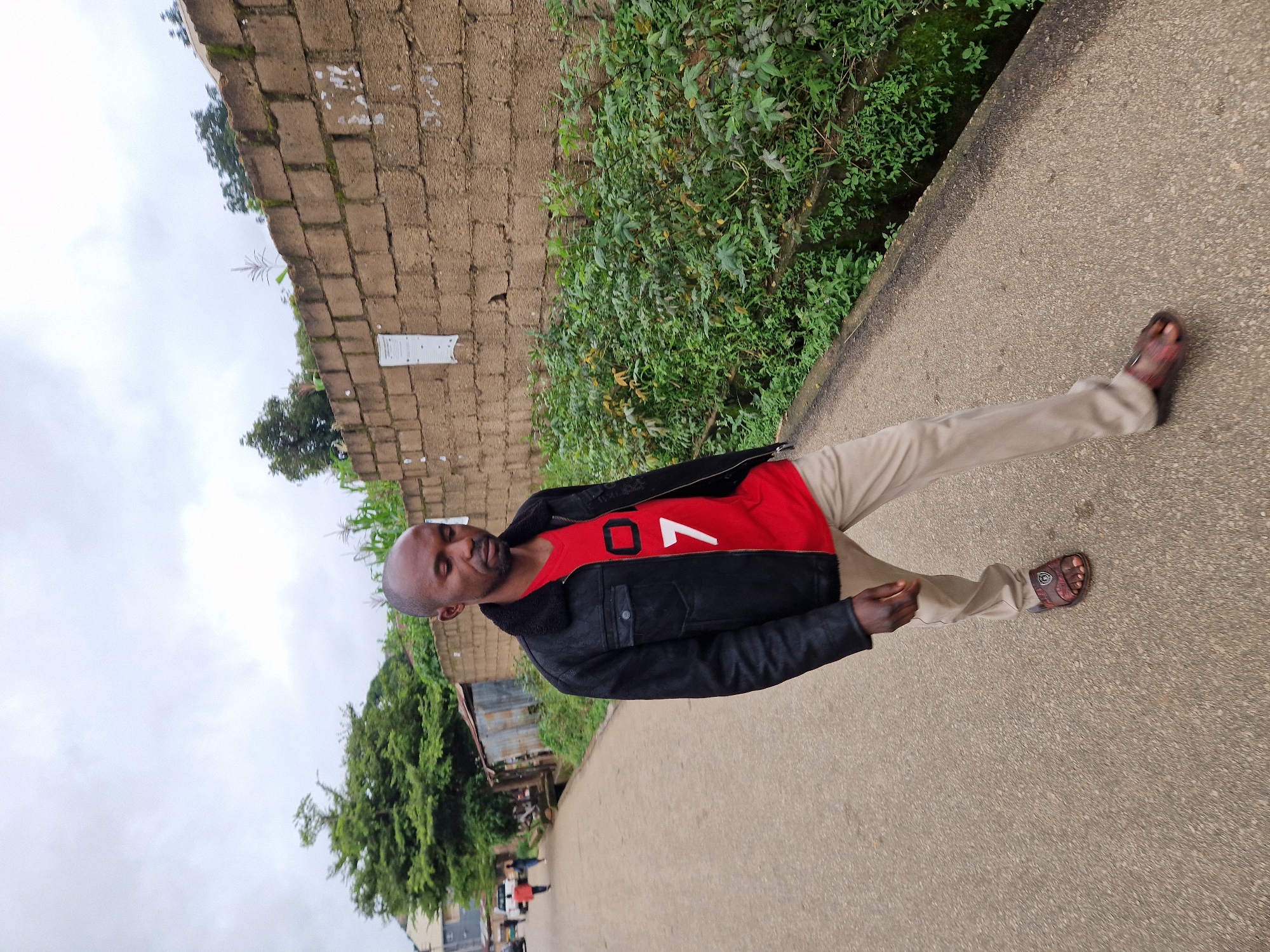 In Kabong, Nigeria, a man walks on an asphalt road with a brick wall to his right and lush green vegetation growing in a ditch alongside the wall. He is wearing a black jacket with a furry collar over a red t-shirt with a white design featuring the number "7". He also wears light-colored pants and brown sandals. In the background, a few people and vehicles are visible on the road, along with some zinc-roofed structures and trees. The sky is overcast.