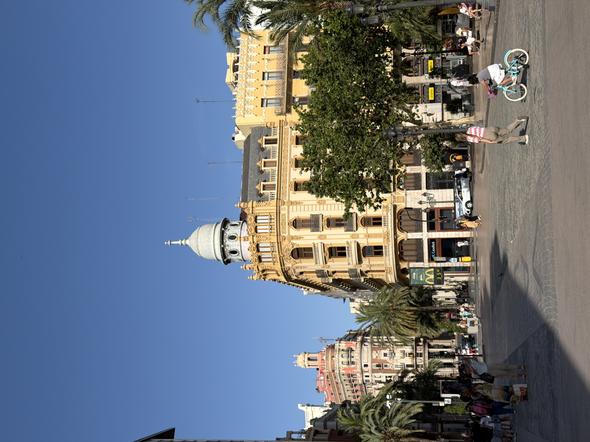 In an urban square under a clear blue sky, a variety of architectural styles are visible. The central focus is a building with ornate yellow facade, balconies, and a distinctive domed cupola. To the left, another building with a pink and white facade stands out, featuring a smaller domed tower. Palm trees are scattered throughout the scene, adding a tropical element to the city environment. Several people are present in the square. On the right side, a young person rides a light blue bicycle. In the mid-ground, a woman in a striped shirt and olive pants walks away from the camera, while another woman in a patterned dress takes a photo with her phone. Other individuals are seen walking, sitting on benches, and interacting in various parts of the square. Several businesses are visible, including a McDonald's with a digital display showing "35°C" and a CaixaBank. Cars and a street light are also present. The ground is paved with cobblestones.