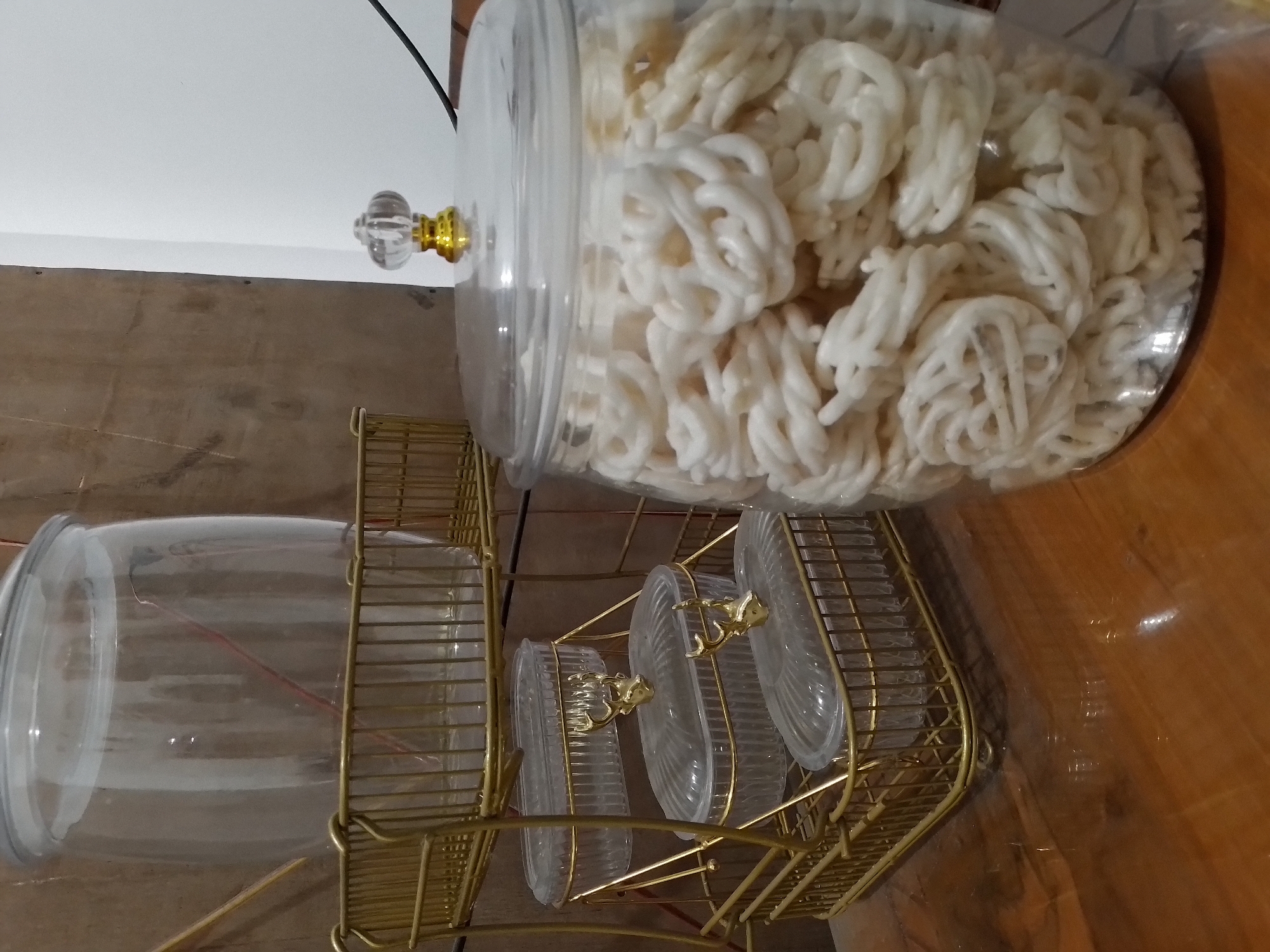 In Ngancar, Indonesia, a wooden table is laden with clear glass jars and a gold tiered stand filled with food items. The stand holds three oval-shaped containers with gold deer-head clasps, each topped with a clear lid. Above these, a larger clear jar sits on the top tier, its contents obscured by the opaque jar itself. To the right, a substantial clear glass jar overflows with white, ring-shaped crackers, its lid adorned with a decorative gold and clear knob. The background is a muted tapestry of textures: a rough, brown wooden panel on the left and a stark white wall on the right. The lighting is dim, casting a warm glow on the polished wooden surface.