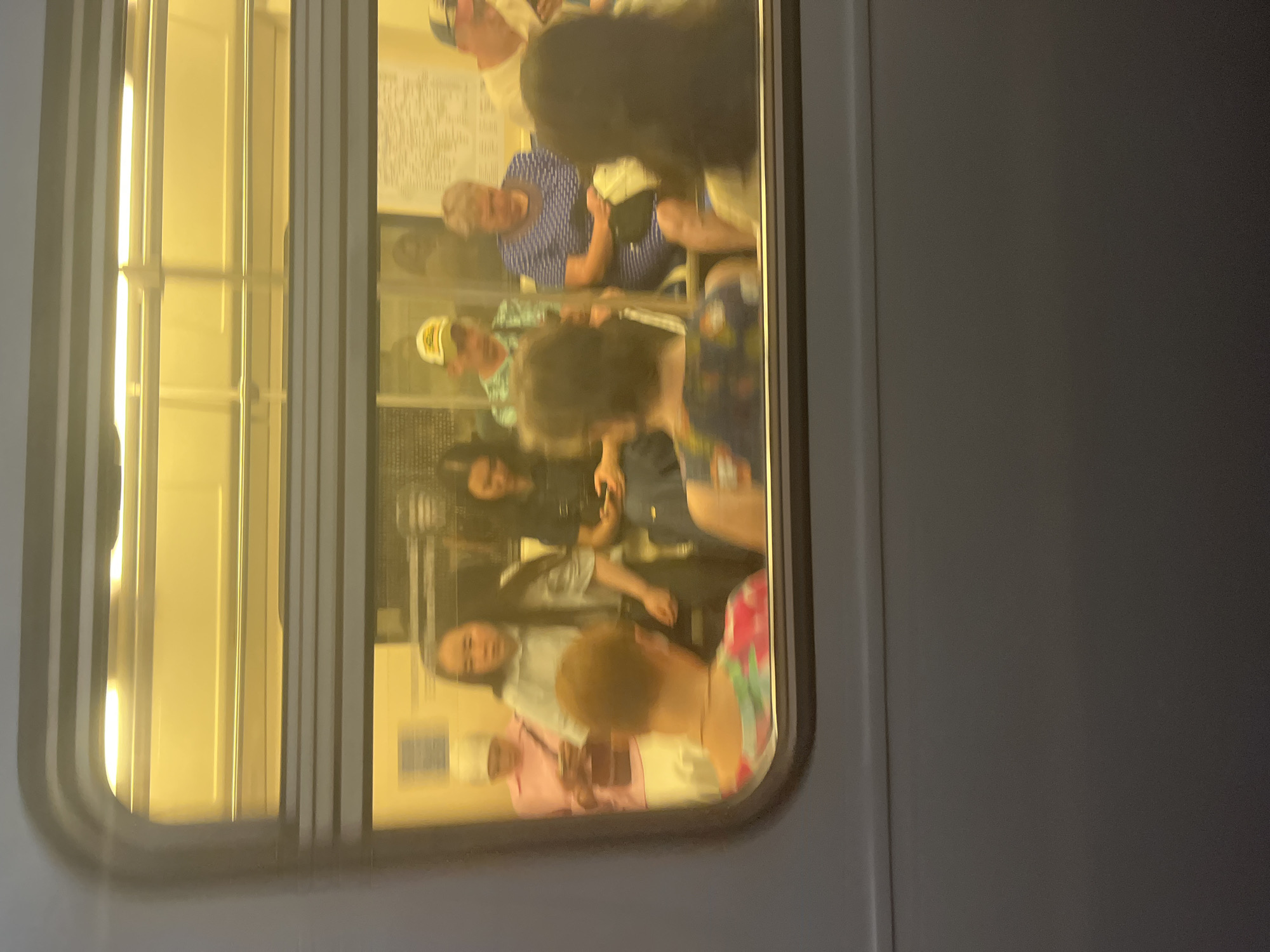 This is a medium shot, taken from outside a subway car, looking through the window at passengers inside. The interior of the subway car is illuminated by bright, yellow overhead lights, creating a warm glow. The window is reflective, partially obscuring the view of the passengers.
There are at least seven people visible, seated and standing. Some are looking down at their phones, while others appear to be looking out the window or at their fellow passengers. Their faces are indistinct due to the motion blur and low resolution of the image. Clothing varies, with a mix of casual attire, including a pink shirt, a light blue shirt, a blue and white patterned dress, and a green and white patterned shirt. One person wears a yellow baseball cap.
The environment appears to be a subway car in Moscow, Russia, suggested by the context of the request, though specific visual cues for this location are not evident within the image itself. The overall impression is one of everyday commuting, with individuals absorbed in their own activities within the shared space of the public transport.