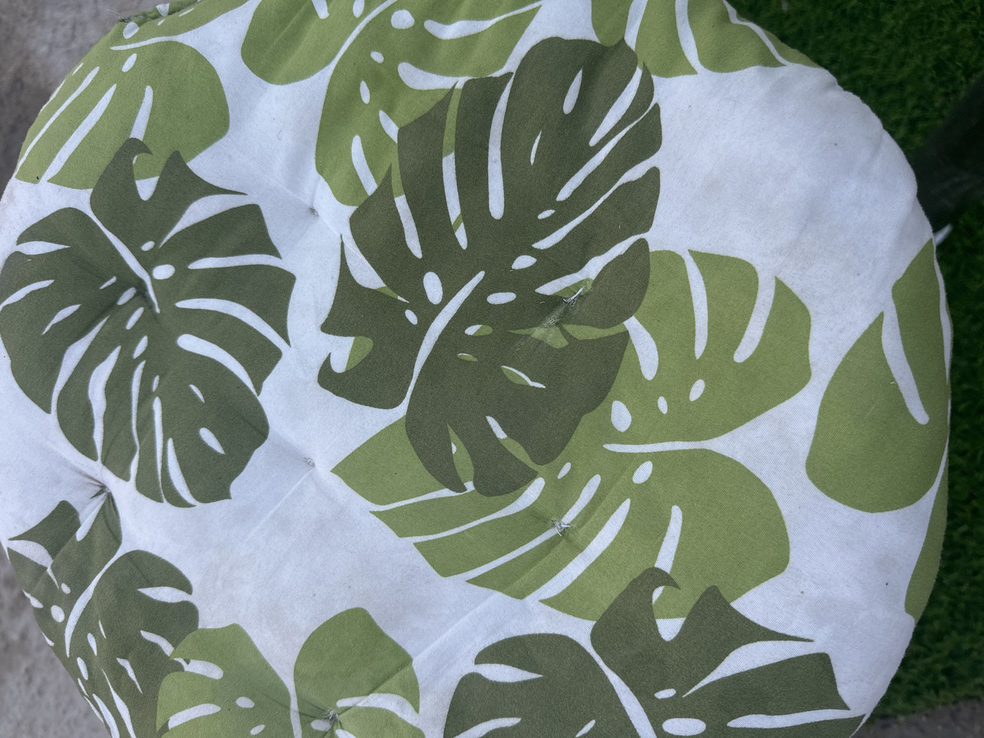 The image shows a close-up, overhead view of a round cushion with a tropical leaf pattern. The cushion is upholstered in white fabric with large green monstera leaves. The leaves are depicted in two shades of green, a lighter green and a darker, olive green, with white cutouts to represent the natural holes in the leaves. The cushion appears to be tufted, with visible stitching creating indentations on the surface. The cushion is placed on what looks like a grey, textured surface, possibly concrete or stone, and a patch of green artificial grass is visible at the bottom right edge of the frame. The scene is outdoors, but the specific location within Atameken, Kazakhstan is not discernible from the image. There are no people, actions, or interactions visible.