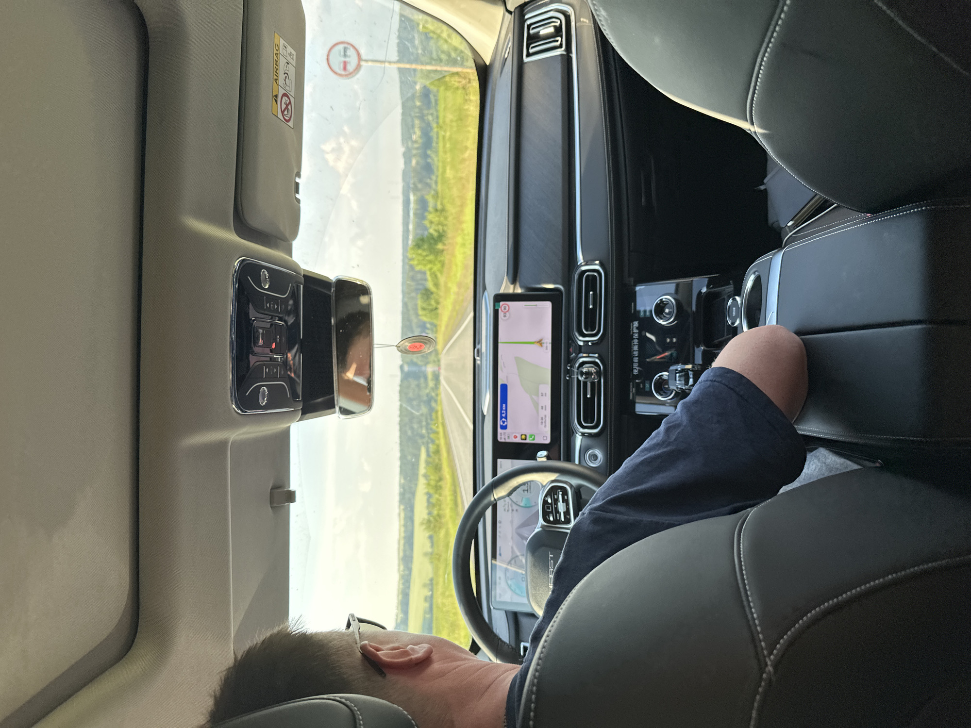 The image shows the interior of a car, likely in Russia, with a person driving. The driver, a man with glasses and light brown hair, is seen from behind, leaning his head against the headrest. He is wearing a navy blue t-shirt. The car's interior features black leather seats with white stitching and a modern dashboard with multiple screens. The driver's side is on the left, and the steering wheel is visible, displaying a navigation system with a map.
Outside the car, a rural landscape is visible through the windshield, with green fields and trees under a bright, possibly overcast sky. A road stretches ahead, bordered by fields. A traffic sign with a red circle and a horizontal bar is visible on the right side of the windshield, indicating a prohibition.
The lighting suggests it is daytime, likely late morning or afternoon, given the brightness of the sky and the outdoor scene. The overall mood appears calm, possibly indicating a leisurely drive through the countryside. No specific emotions are overtly displayed by the driver, but the relaxed posture suggests a comfortable journey. The visible text includes "AIRBAG" on a sun visor, and various labels and icons on the car's dashboard screens, including navigation information.