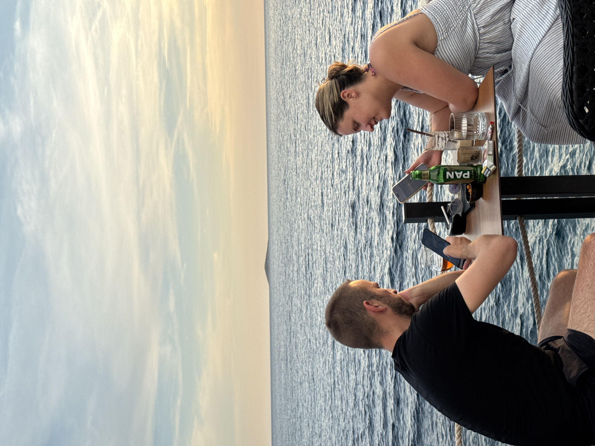 Two people, a man and a woman, are seated at a table by the sea at sunset. The man, on the left, wears a black t-shirt and shorts and looks down at his phone. The woman, on the right, wears a striped dress and is also looking at her phone, smiling. On the table are drinks, cigarettes, and a small bowl of nuts. In the background, the sea stretches to the horizon, with a small island visible in the distance. The sky is a pale blue with wispy clouds, tinged with the warm colors of the setting sun. This scene is likely located in Primošten, Croatia, a coastal town known for its picturesque sunsets over the Adriatic Sea.