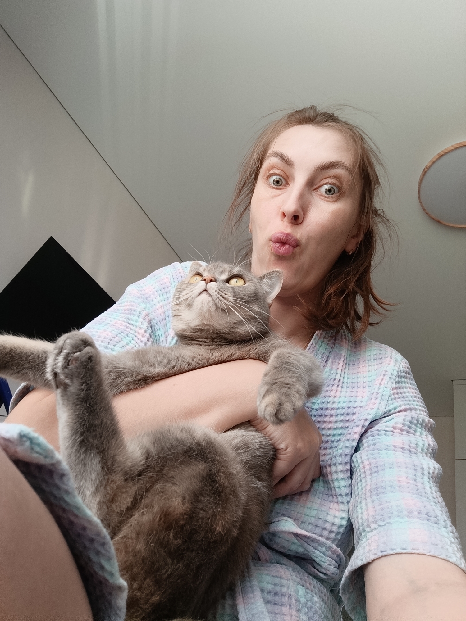 A woman is holding a gray cat in her arms. The woman has auburn hair pulled back, and she is wearing a waffle-patterned robe. Her eyes are wide and she has puckered lips. The cat has yellow eyes and outstretched paws. The location is indoors, in a room with white walls and an oval mirror on the wall. A dark, rectangular object is mounted on the wall. The location is Chaikovsky, Russia.
