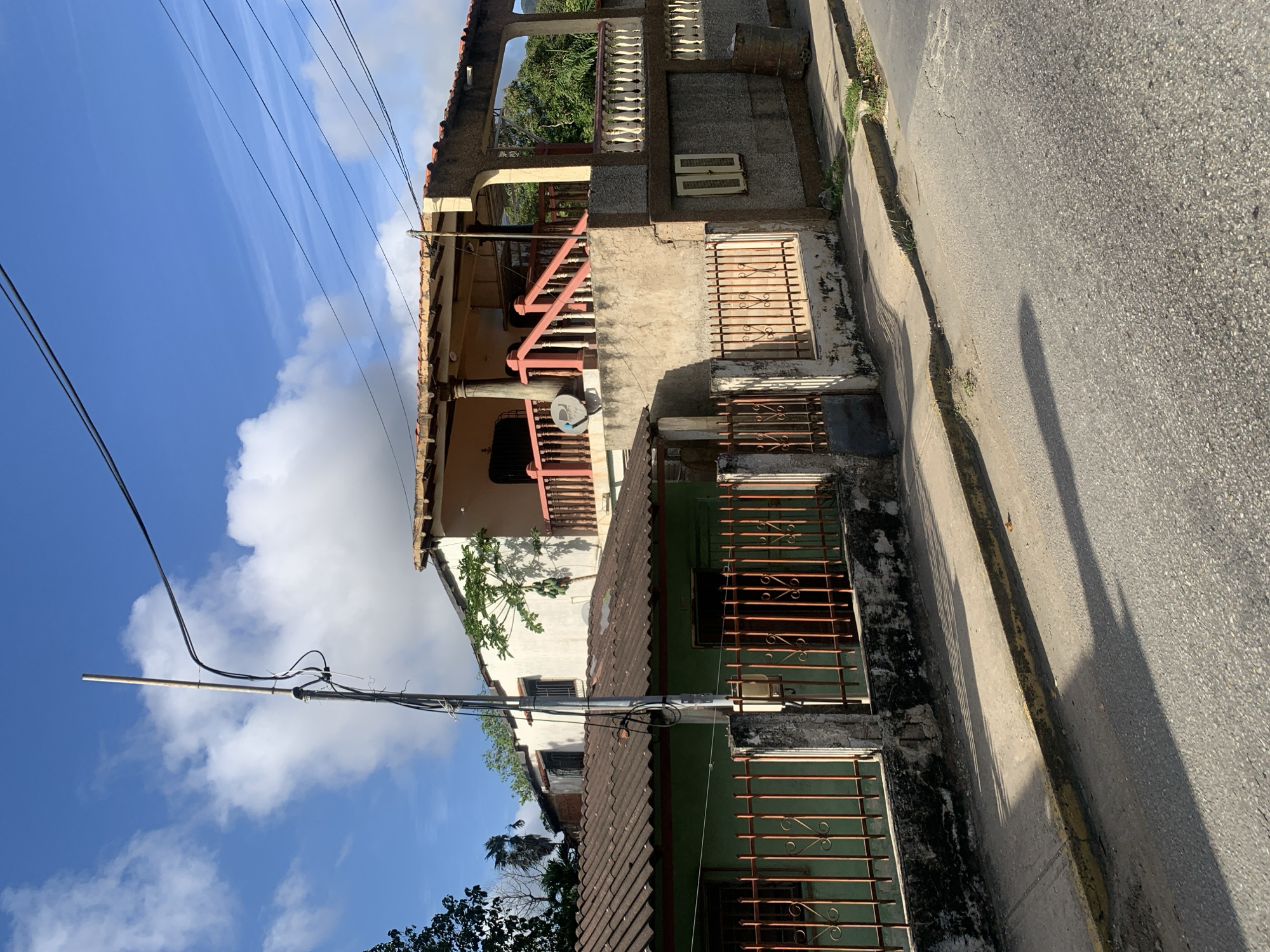 The image shows a street scene in La Asuncion, Venezuela, with two attached residential buildings and a concrete sidewalk. The foreground is dominated by a paved street with a yellow-lined curb. To the left of the street is a concrete sidewalk, casting long shadows from an unseen light source. Behind the sidewalk are two houses. The house on the left has a green-painted exterior and a corrugated metal roof. It is bordered by an orange metal fence with decorative elements. The house on the right is larger and appears to be two stories. It has a light-colored stucco exterior and a tiled roof. This building features a covered balcony with a pink railing and white-capped columns. A satellite dish is attached to the upper section of this house. A tall utility pole with several electrical wires runs vertically through the image, near the center. In the background, lush green trees and vegetation are visible. The sky is blue with scattered white clouds. No people are visible in the scene.