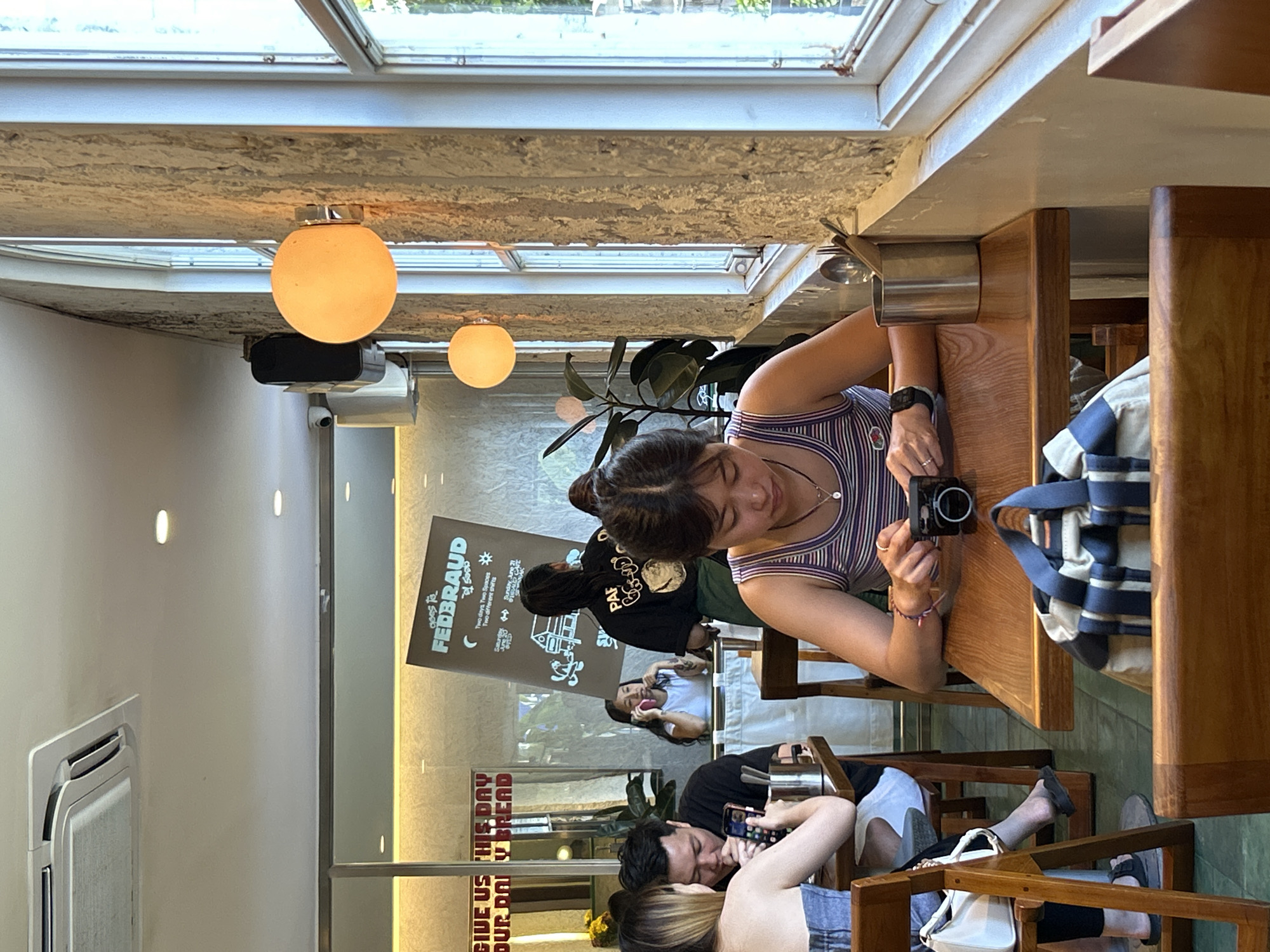 The image depicts the interior of a cafe in North Kuta, Indonesia. The focus is on a young woman in the foreground, seated at a wooden table, holding a small black camera. She is wearing a striped tank top. In front of her on the table is a blue and white striped duffel bag.
Behind her, another woman is visible in the middle ground, on a phone call. She is wearing a white t-shirt and has tattoos on her arms. To her left, a man in a black t-shirt is also looking at his phone. A woman with blonde hair is seated next to him.
The cafe's decor includes a textured, light-colored wall with a rough finish, large windows with white frames, and two spherical pendant lights emitting a warm glow. A sign with text, partially obscured, is visible in the background. The floor appears to be tiled. Other patrons and tables are present in the cafe, suggesting a busy atmosphere.