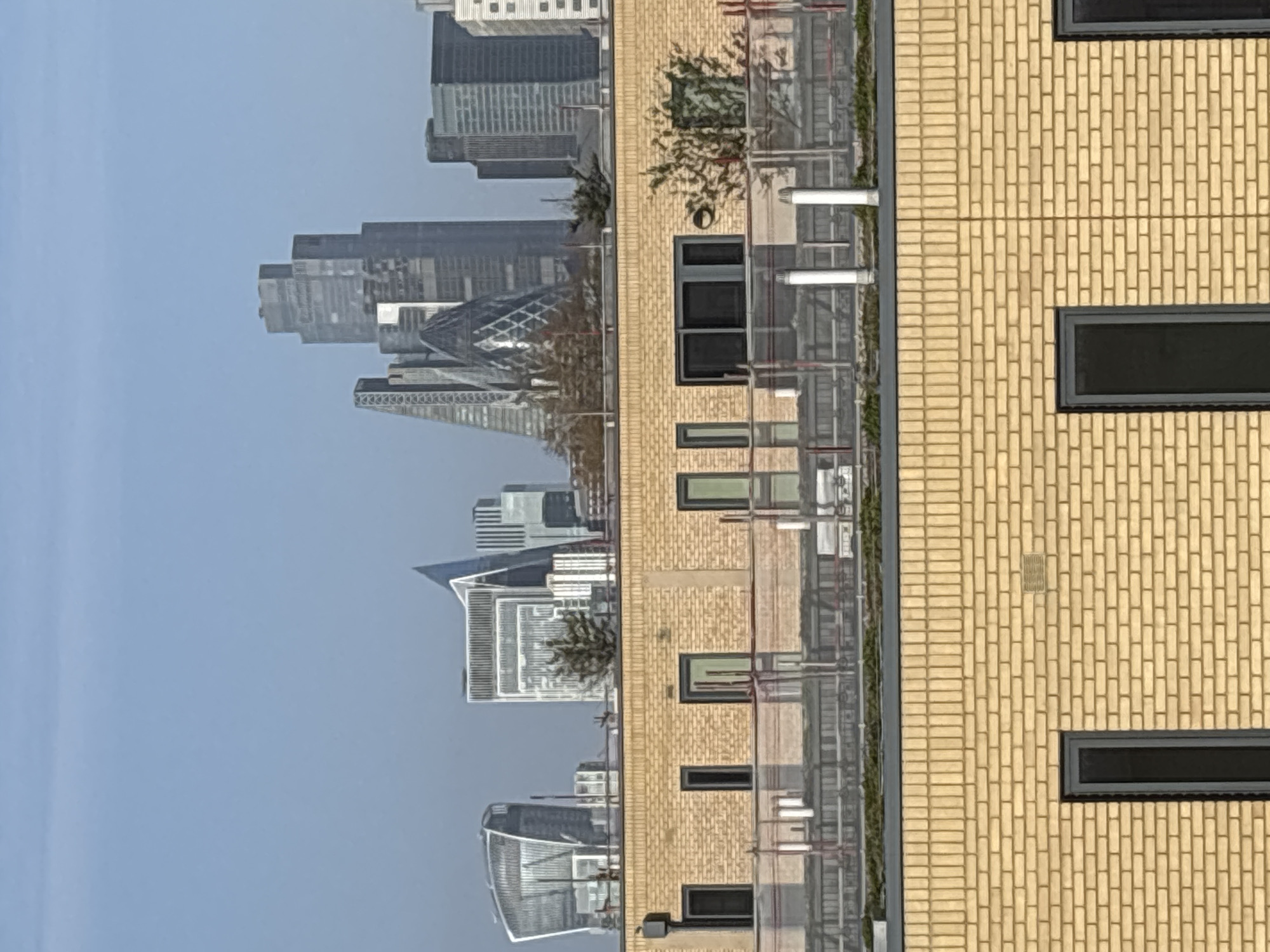 The image shows a partial view of the London skyline, featuring the Gherkin building and other skyscrapers, seen through the window or from the balcony of a brick building. The brick building has a yellow facade with dark window frames and a series of balconies with metal railings and white piping. Some greenery is visible on the balconies. The sky is a light blue color. The image is taken from a high angle, looking out toward the city.