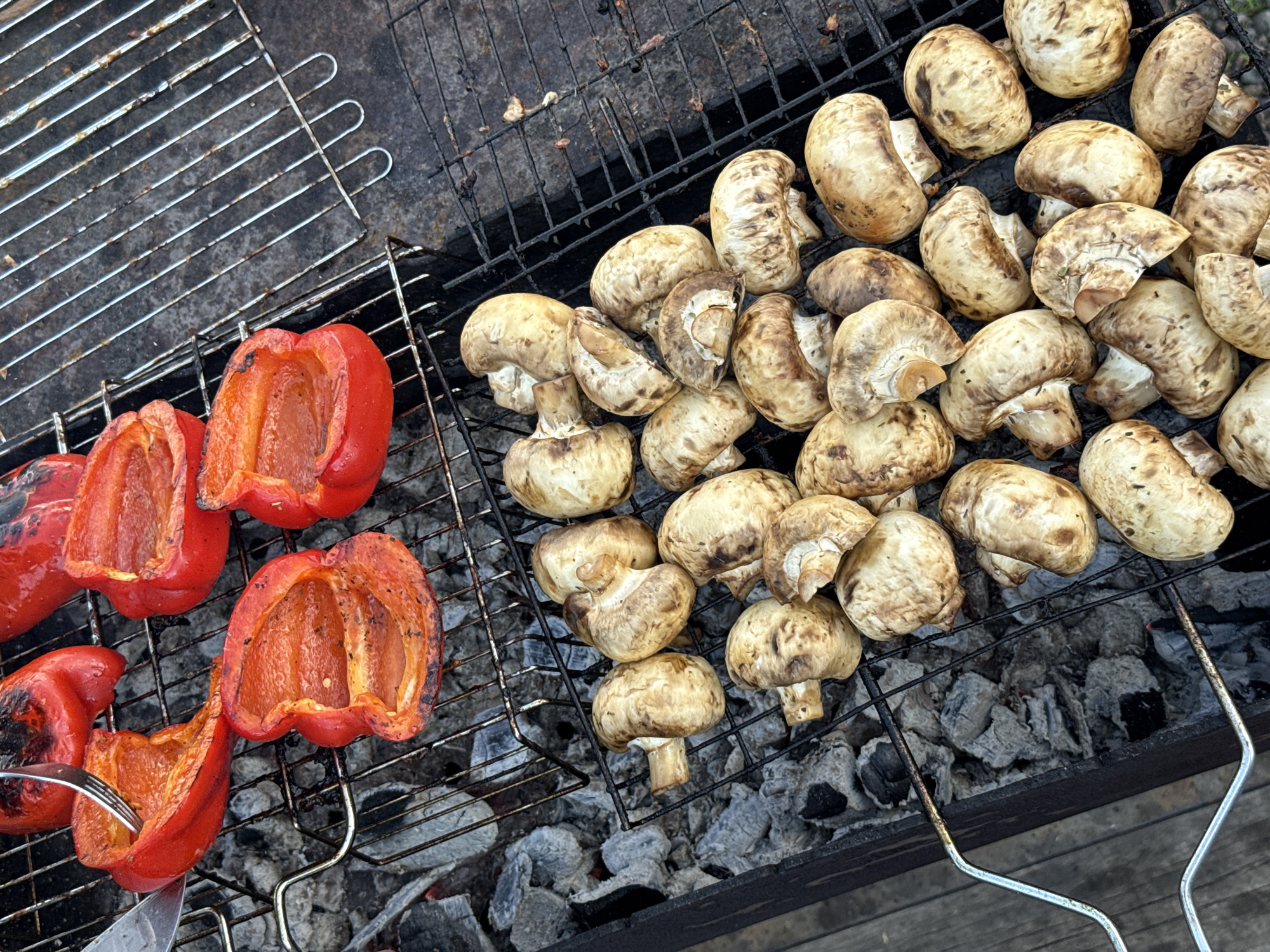 The image shows red bell peppers and whole button mushrooms grilling on a charcoal grill. The grill sits above grey charcoal. A fork is inserted into one of the pepper halves. The location is not discernable from the image.