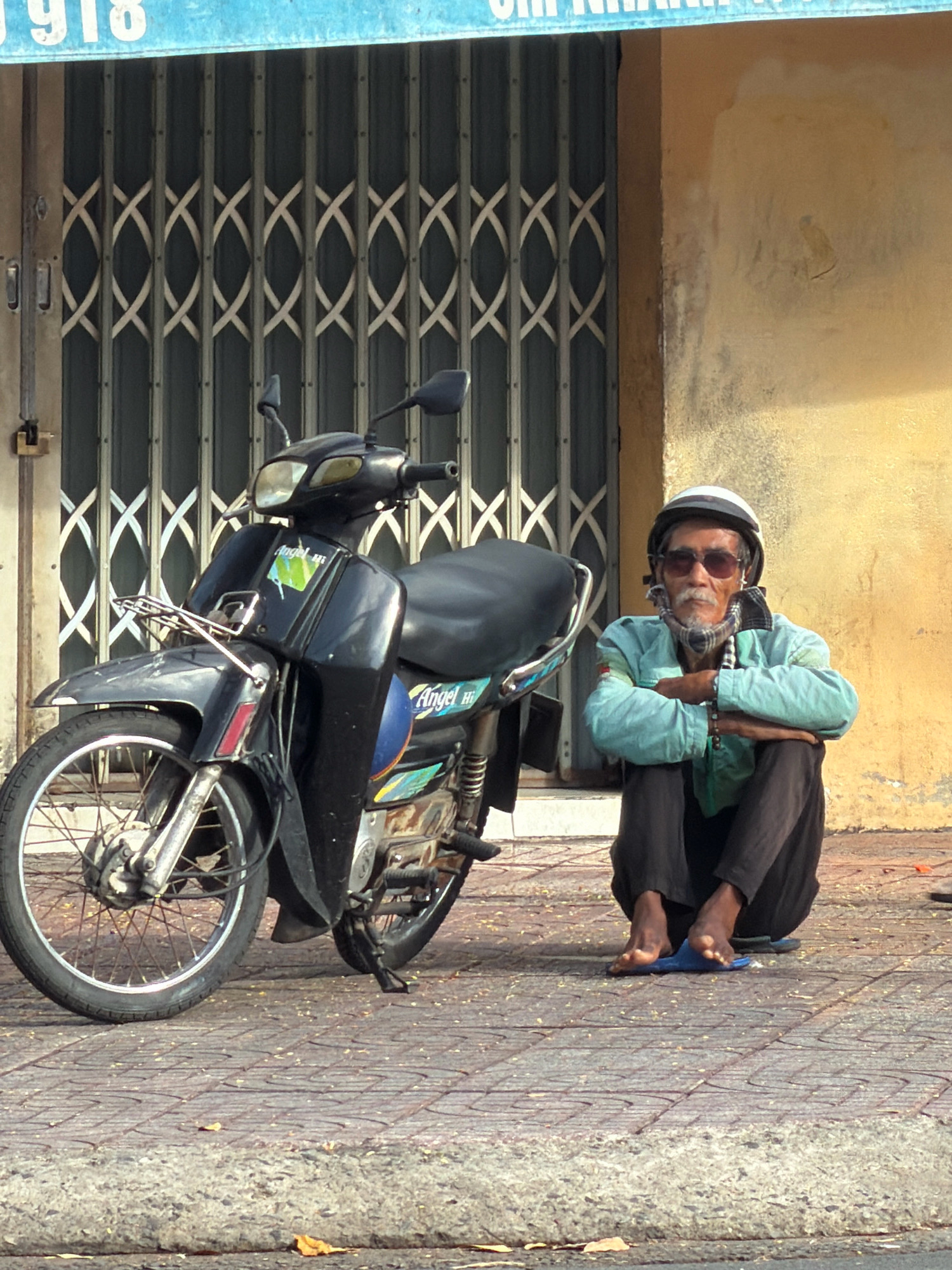An elderly man with a white helmet, sunglasses, and a scarf around his neck sits cross-legged on a tiled sidewalk in Nha Trang, Vietnam. He is wearing a light blue jacket and dark pants. Next to him is a black motorcycle. Behind them is a building with a yellow wall and a metal grate door. A blue sign with white numbers and letters is visible above the grate. The man's feet are bare, and he is wearing blue flip-flops. The scene appears to be in an urban area, possibly a street or shopfront.