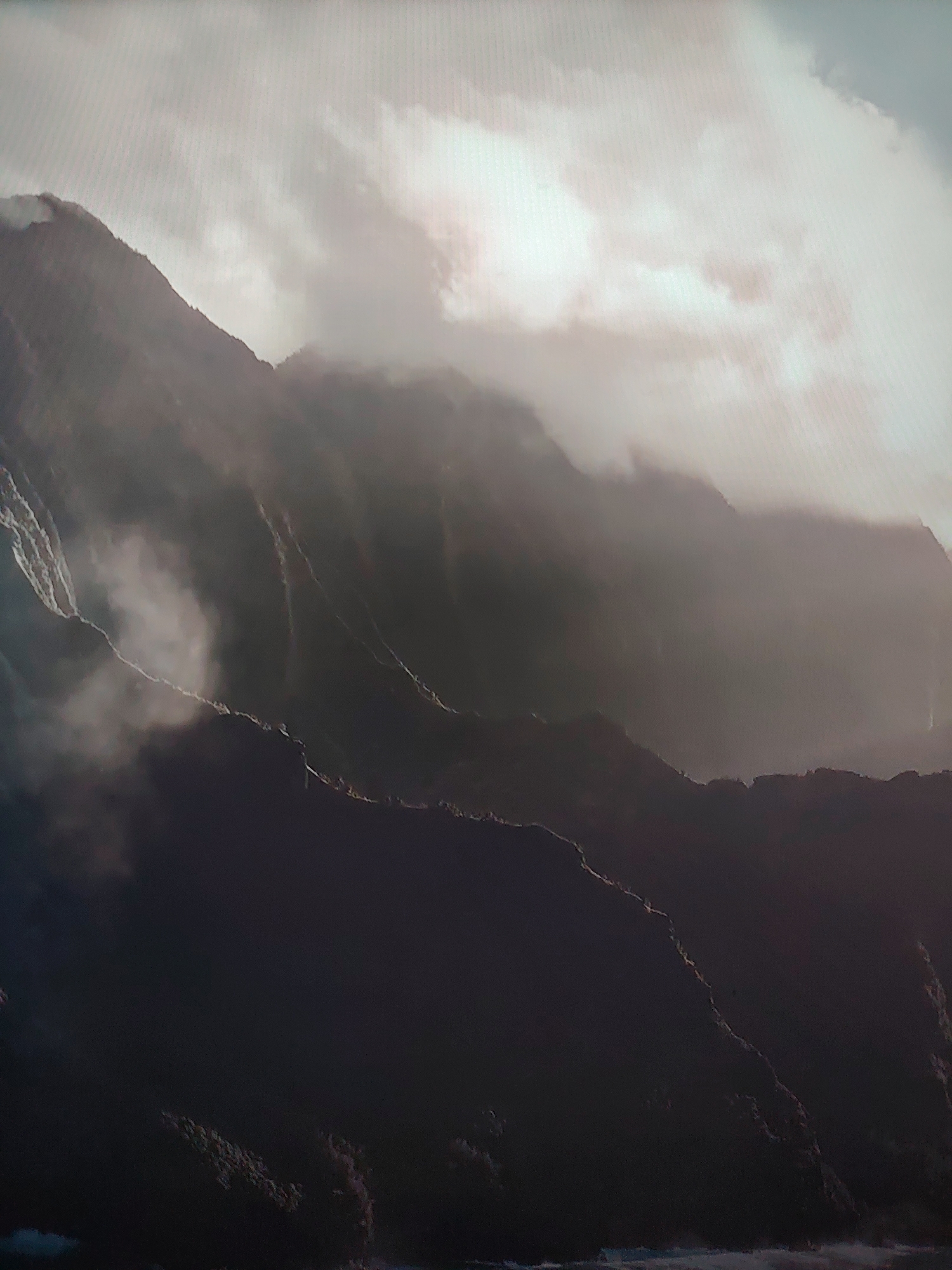 The image depicts a mountain range shrouded in fog and clouds. The scene is characterized by varying shades of grey, ranging from the dark, shadowed mountain slopes in the foreground to the brighter, cloud-covered sky above. Water can be seen on the left side. The location is Unknown, Unknown.