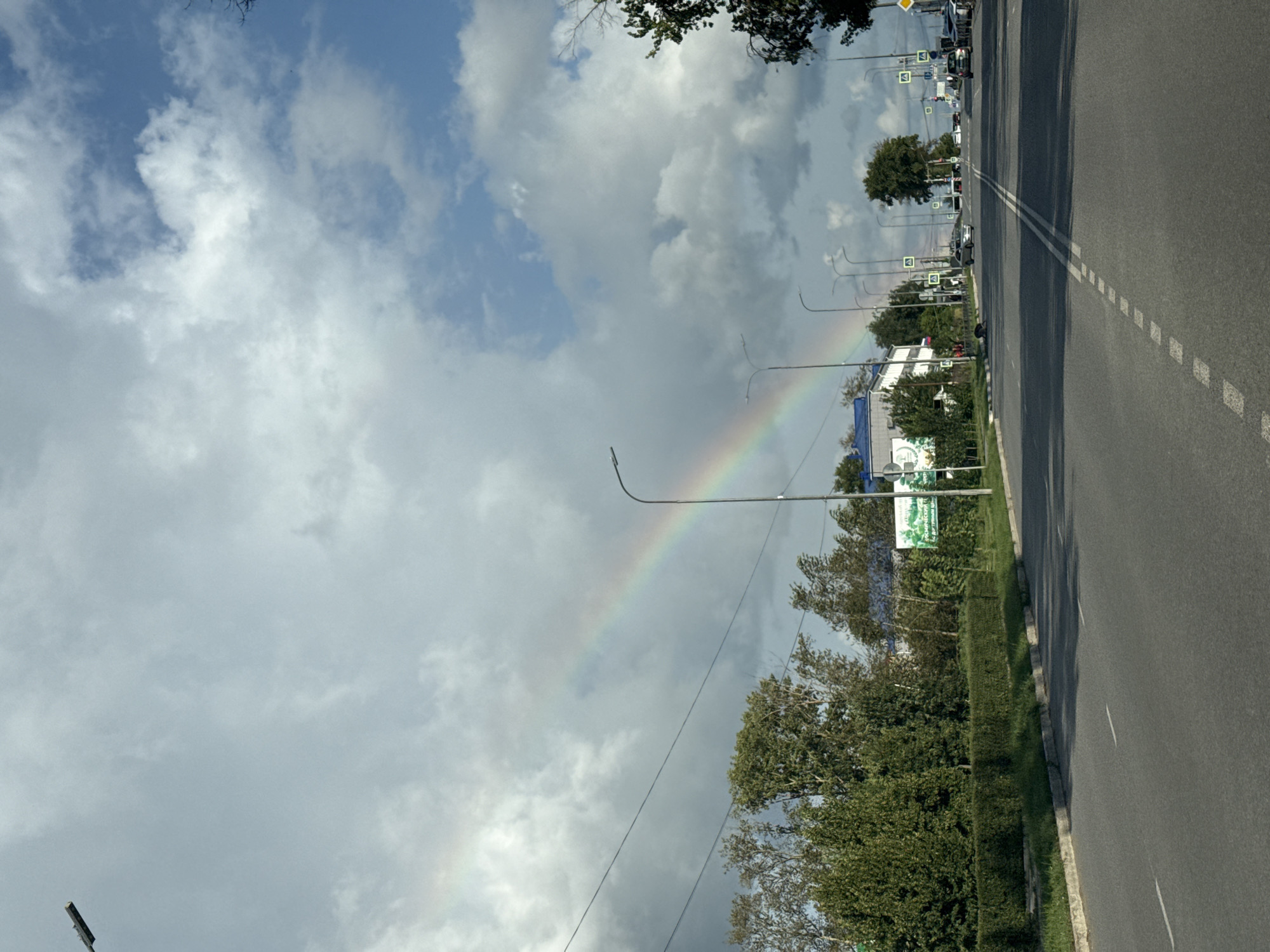 This is a wide-angle, eye-level, vertical shot of a road in Chaikovsky, Russia, featuring a rainbow stretching across a cloudy sky. The foreground is dominated by an asphalt road with white dashed lines indicating lanes. On the left, a neatly trimmed green hedge lines the roadside, followed by a row of lush green trees. Behind the trees, a modern white building with a blue roof is visible. Several streetlights are positioned along the road. Further down the road, traffic signs and other vehicles can be seen. The sky is a mix of blue and dramatic grey clouds, with a prominent rainbow arcing from the left side of the frame towards the right. The overall scene depicts a typical urban or suburban environment under a weather-affected sky.