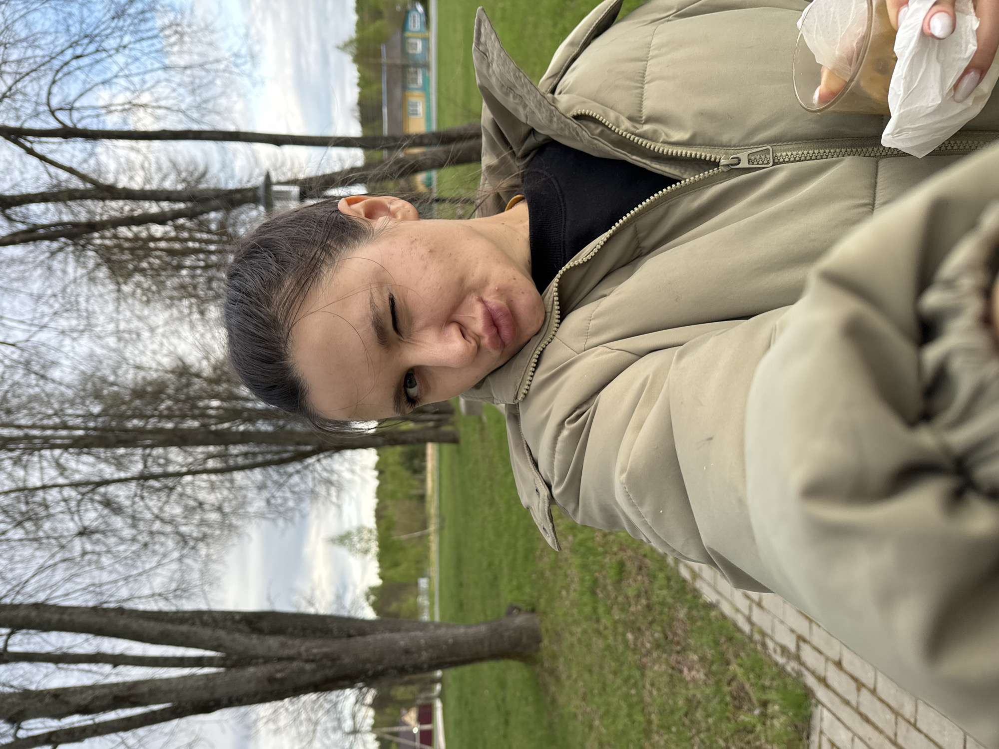 A selfie taken outdoors in Красный Маяк, Russia, shows a person wearing a beige puffer jacket. The individual is holding a clear glass with a white tissue or napkin in their right hand. Their expression is a slight smirk or pucker. The background features a grassy area with trees and a building in the distance. The ground near the person appears to be paved.