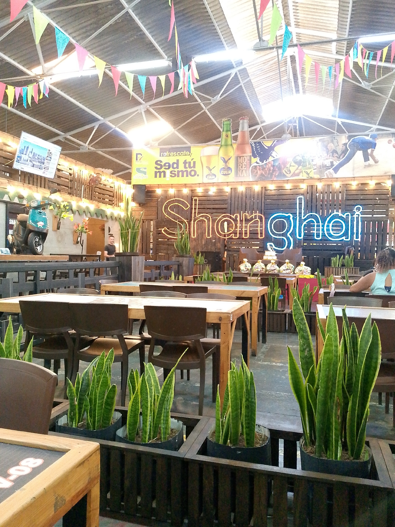 The image depicts an indoor restaurant in an unidentified city. Tables and chairs are arranged throughout the space, many surrounded by planter boxes containing snake plants. In the background, a wooden wall is decorated with the name "Shanghai" in neon lights, a billboard featuring beer advertisements, and a small television screen. Bunting hangs from the corrugated metal ceiling, adding to the festive atmosphere. A scooter is visible on a raised platform to the left, adding a unique decorative element. Several individuals are seated at tables, engaged in conversation or dining.
