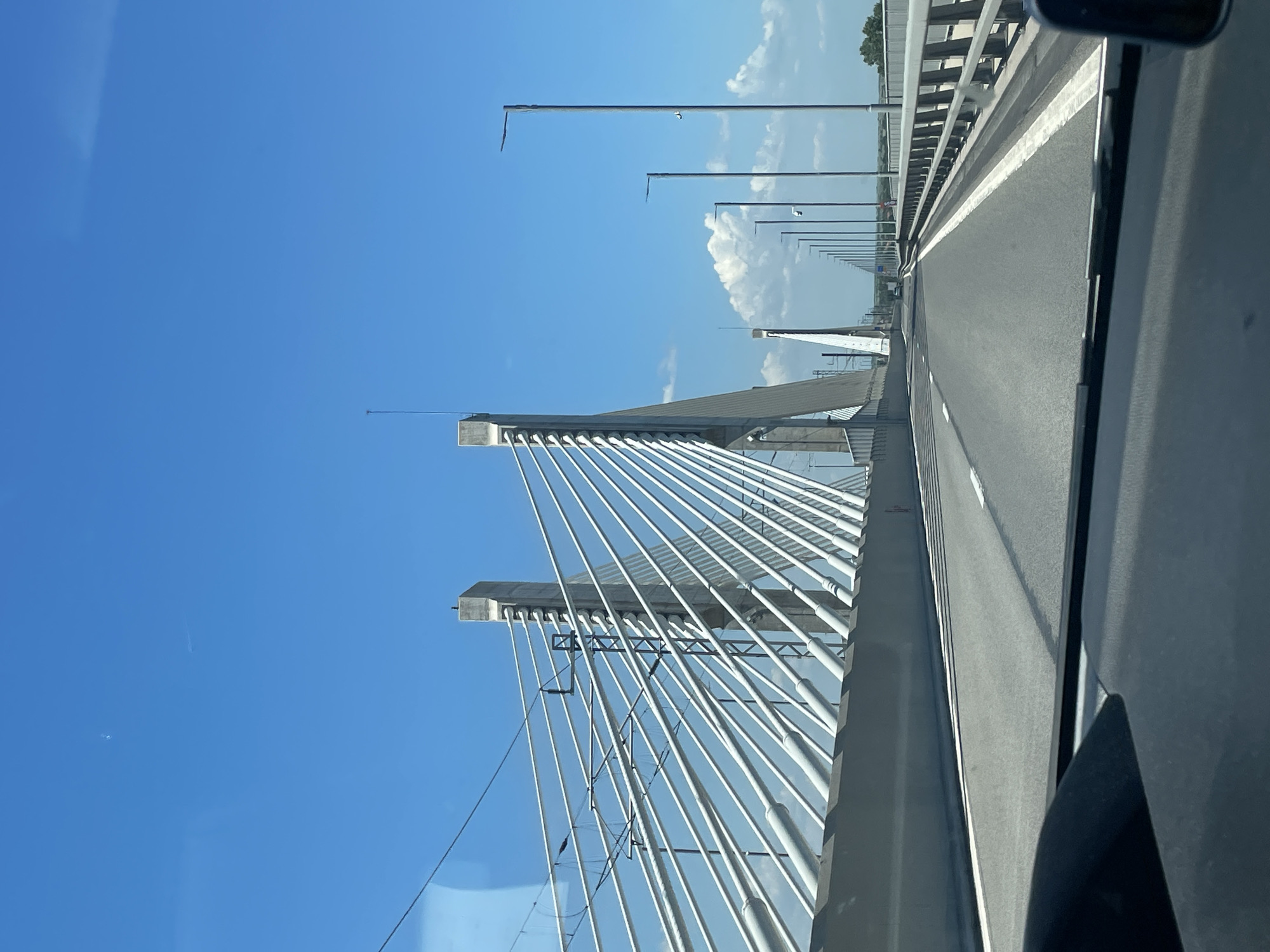The image depicts a straight-ahead view from the interior of a vehicle traveling on a multi-lane cable-stayed bridge in Antimovo, Bulgaria. The road surface is asphalt, with clear white lane markings and a solid white line defining the right shoulder. Dark gray concrete barriers line both sides of the roadway.
On the left side, two prominent, light gray concrete pylons of the cable-stayed bridge are visible, from which numerous white-sleeved tension cables fan out to support the bridge deck. Overhead, an intricate network of dark gray wires and support structures, consistent with an electrified railway catenary system, parallels the road next to the pylons.
To the right, a series of modern, dark gray streetlights recede into the distance. The nearest streetlight on the right features a small white surveillance camera or sensor. Beyond the bridge's end, a flat, green landscape is faintly visible under a bright, clear blue sky dotted with scattered white cumulus clouds. The lower edge of the vehicle's dark dashboard or windshield frame is visible at the bottom of the image. No individuals or other vehicles are distinctly visible, aside from a highly indistinct shape in the far distance on the right side of the bridge. The scene is illuminated by strong daylight.