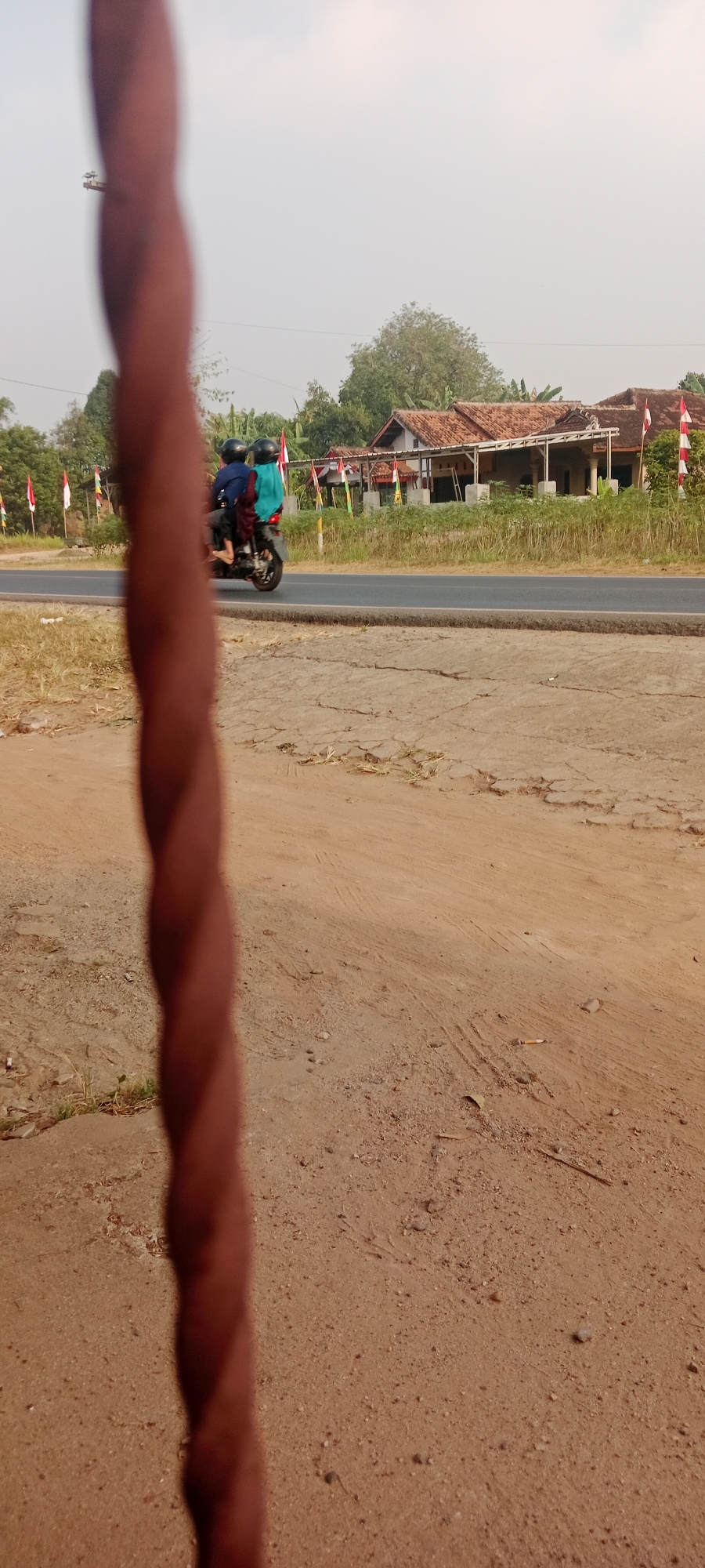 In the foreground of the image, a vertically oriented, twisted reddish-brown object dominates, out of focus, with dry, cracked brown earth visible behind it, sparsely dotted with small stones and dry vegetation. Beyond the earth, a paved road runs horizontally across the midground. On the road, a motorcycle is traveling from left to right, carrying two individuals. The person in the rear is wearing a turquoise head covering and dark clothing. The person in the front is wearing a black helmet and dark clothing. The motorcycle appears to be dark in color. Flanking the road are grassy verges and scattered trees. Behind the verge on the right, a series of houses with reddish-brown tiled roofs are visible. Red and white flags are displayed along the front of these houses. The sky above is overcast and hazy. The overall scene suggests a rural or semi-rural setting in Gondang Rejo, Indonesia, likely on a road with houses and vegetation.