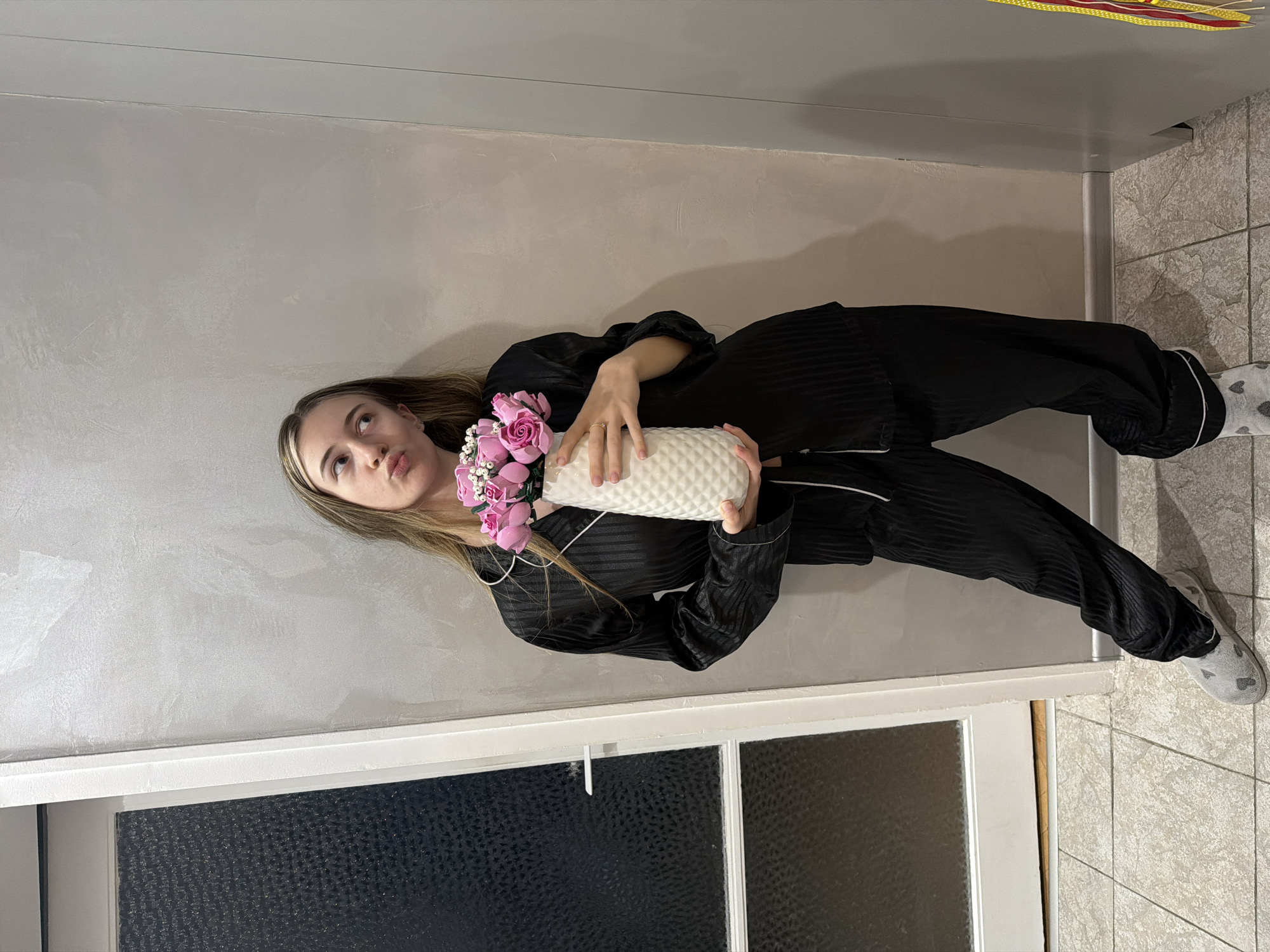 A young woman wearing a black satin pajama set stands indoors. She is holding a white vase filled with pink roses. The background includes a light grey wall, a dark window, and tiled flooring. The location is Plovdiv, Bulgaria.