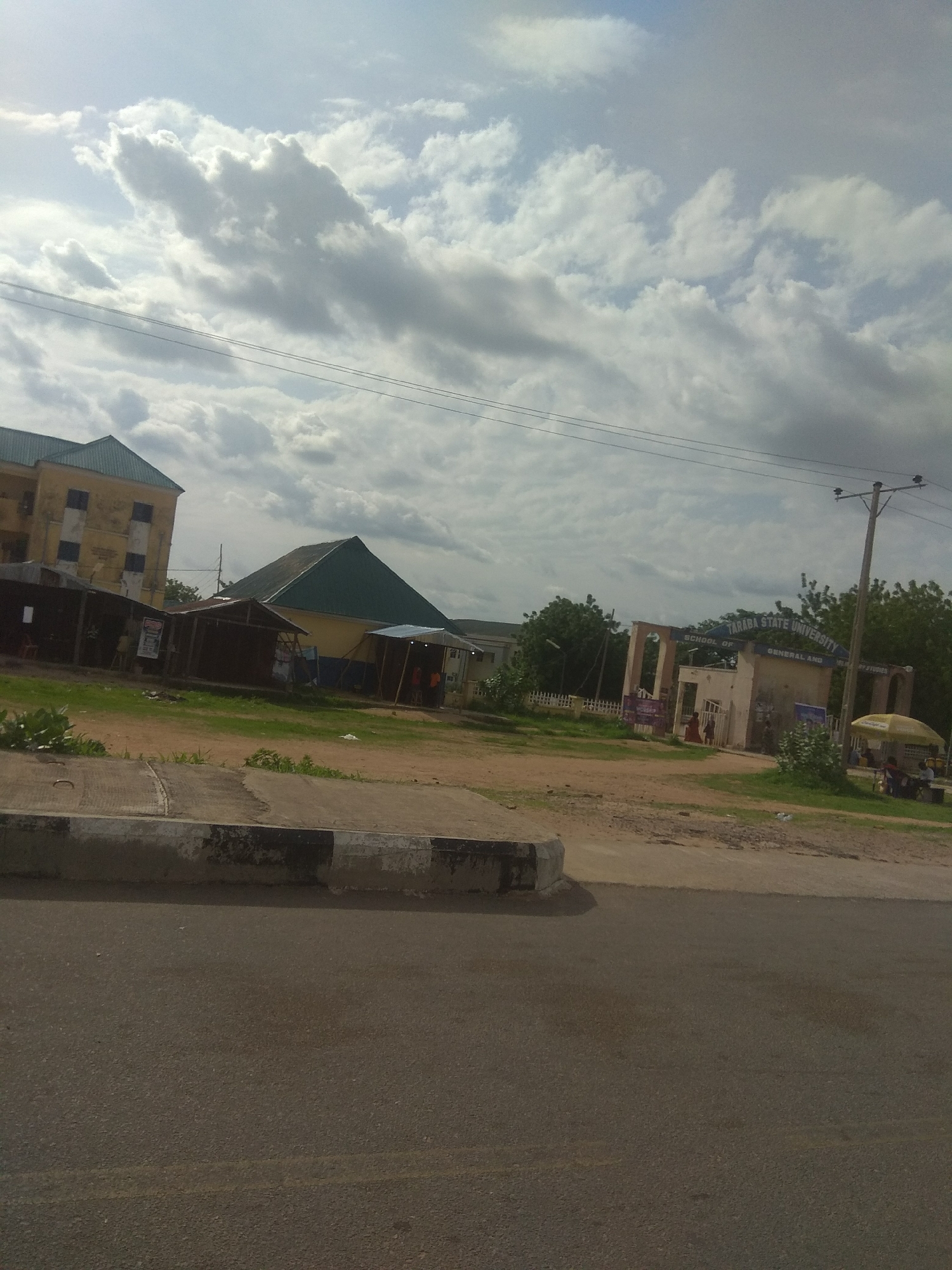 The image shows an overcast sky with scattered clouds. In the foreground, a paved road is visible with a concrete curb. Behind the curb, there is a patch of dry grass and dirt. To the left, a multi-story building with a yellow facade and green roof is partially visible. Adjacent to this building, several small, open-fronted stalls with corrugated iron roofs stand in a row.
Further into the scene, a larger building with a prominent green, pitched roof is visible, situated behind the stalls. To the right of this building, there is an entrance gate with the text "TARABA STATE UNIVERSITY SCHOOL OF GENERAL AND CONTINUING STUDIES" displayed above it. The gate itself is constructed with arched openings and weathered beige walls.
Beyond the gate, more greenery and trees can be seen. A utility pole with wires runs across the top portion of the image. In the far right, a yellow umbrella shades a small area, and some figures appear to be gathered beneath it. The overall impression is of a somewhat utilitarian and possibly developing educational institution in a warm climate, likely during the daytime given the brightness of the sky. The presence of the stalls suggests a marketplace or informal commercial activity near the university entrance.