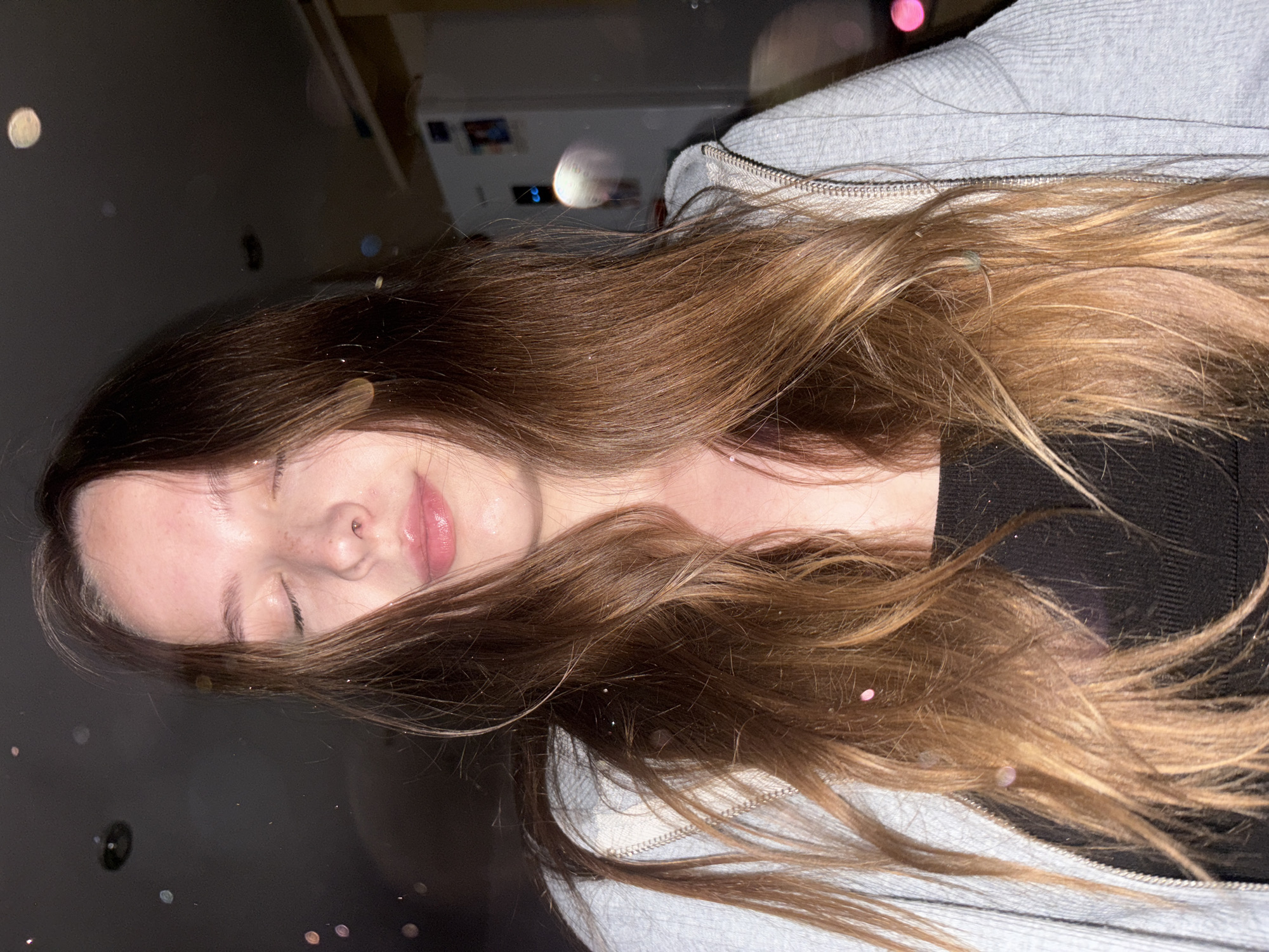 A young woman with closed eyes and light brown hair is pictured in a close-up, waist-up shot. She is wearing a black top and a grey zippered hoodie. Her face is tilted slightly upwards, and her lips are glossy. The background is dark and out of focus, with some blurred lights, suggesting an indoor setting at night. There are small, scattered sparkly specks visible on her hair and clothes. The image does not provide any identifiable details about the location in Chaikovsky, Russia, or any specific environmental context that would suggest it is within that city.