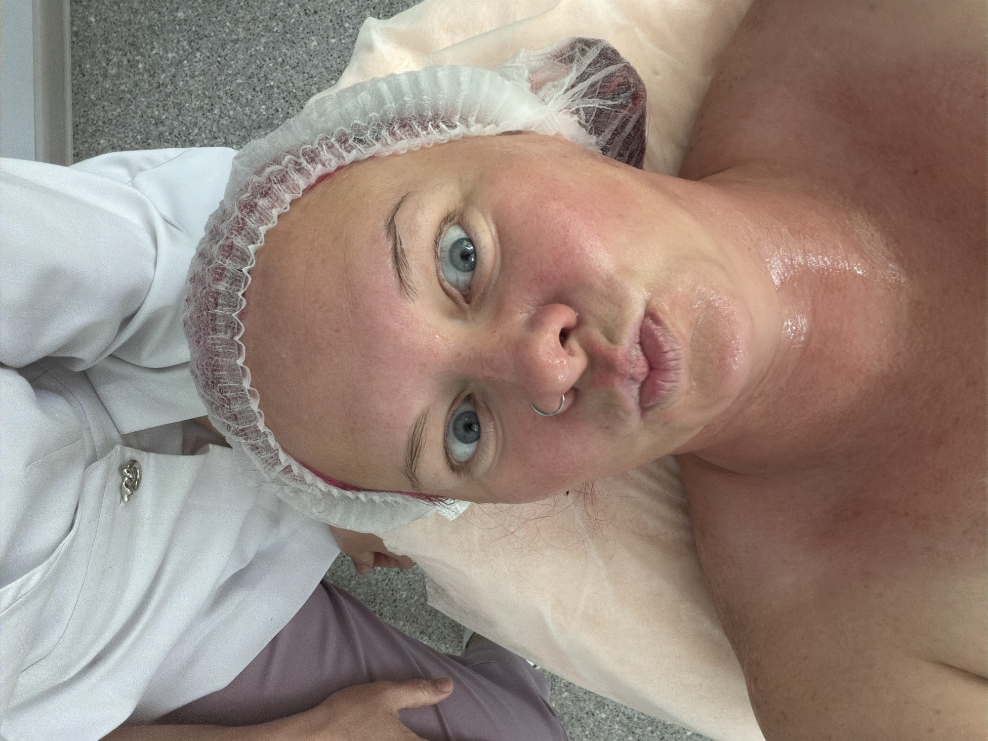 An overhead shot depicts a person, likely a woman, lying on her back, seemingly on a treatment bed covered with a light-colored disposable sheet. She is wearing a white disposable hair net or shower cap that covers her hair. Her face, neck, and upper chest are visible and appear to be covered in a shiny, liquid, or oily substance, suggesting she is undergoing a facial or body treatment. Her blue eyes are wide open, looking directly at the camera, and her lips are slightly pursed. A silver nose ring (septum piercing) is visible.
To the left of the primary subject, another person is partially visible, wearing a white uniform jacket with a distinctive silver clasp or button on the lapel. The background shows a light grey, possibly speckled, floor or wall surface. The setting appears to be an indoor professional environment, such as a beauty salon, spa, or clinic, possibly located in Chaikovsky, Russia, given the provided context. No specific time of day or weather conditions are discernible, and there is no visible text. The overall expression of the subject is one of direct engagement, possibly playful or slightly surprised.