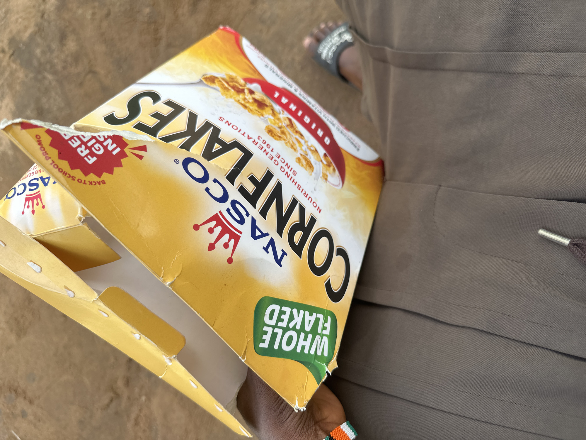 A person in brown pants and sandals is holding an open box of NASCO Cornflakes in what appears to be Ikot Ekpene, Nigeria, as indicated by the product branding. The box is yellow with the brand name and "WHOLE FLAKED" text. The box is open at the top, and its condition suggests prior use. The ground is visible in the background. The person is wearing a beaded bracelet on their wrist.