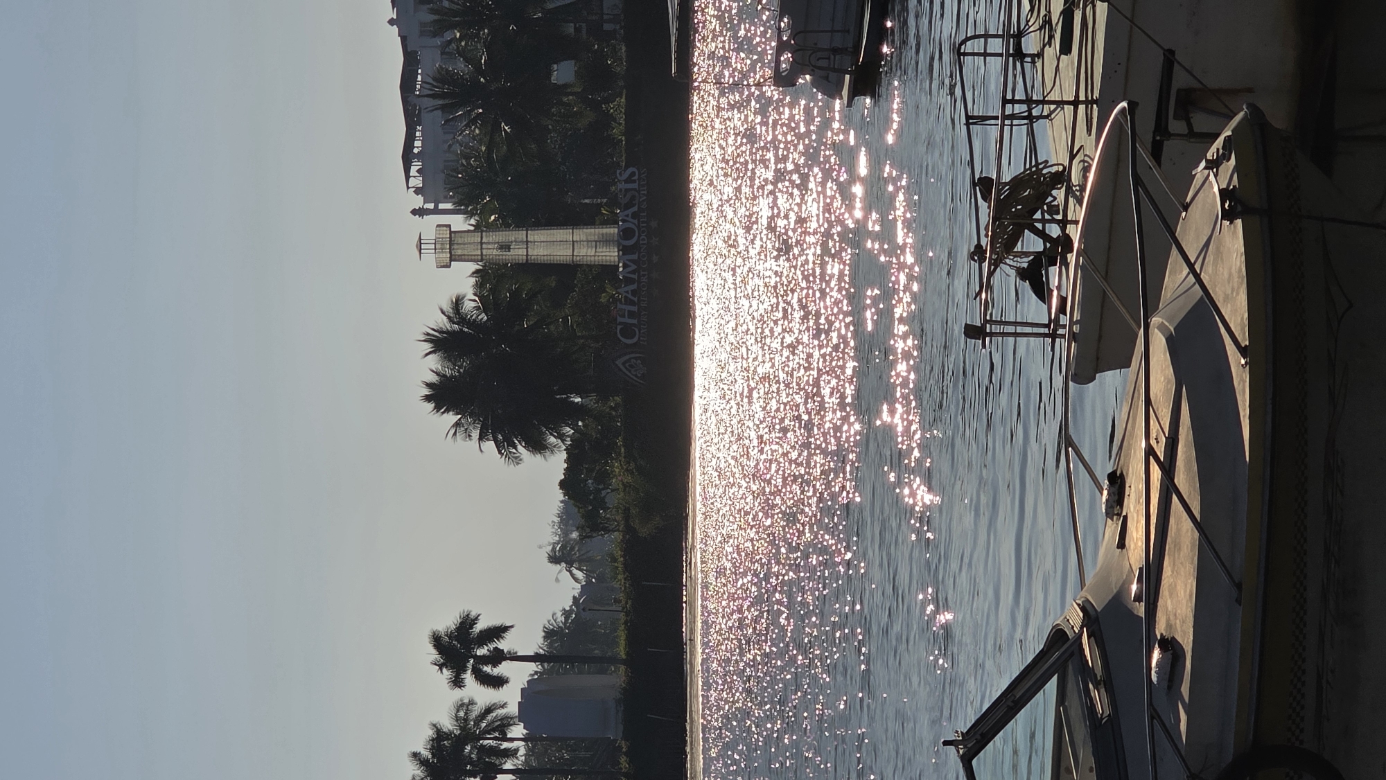 The image depicts a marina scene in what appears to be Bắc Nha Trang, Vietnam. Palm trees and buildings, including a modern lighthouse-like structure, line the shore. A large sign for "CHAM OASIS LUXURY RESORT CONDO & VILLAS" is visible along the shoreline. Sunlight glints off the water's surface. The foreground is dominated by docked boats, with their rigging and hulls partially visible.