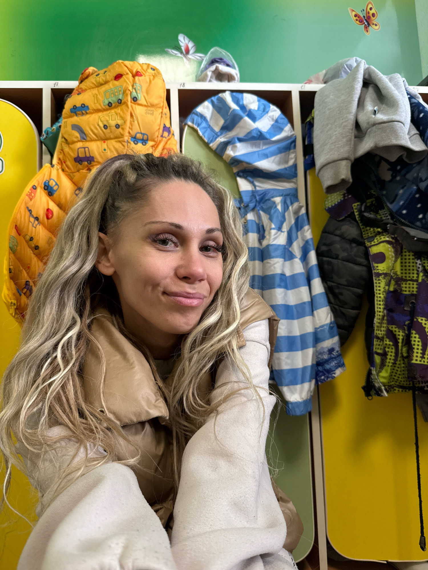 A young Caucasian woman with blonde, wavy hair tied in a high ponytail and wearing a beige puffer vest and a cream-colored sweatshirt poses for a selfie in front of a colorful locker room. She is smiling with dimples visible. Behind her, a row of lockers painted in yellow and green hues are filled with children's outerwear. A yellow puffer jacket with a car print, a blue and white striped jacket, and a gray hooded sweatshirt are hanging in the lockers. A decorative butterfly sticker is on the green wall above the lockers. The scene appears to be in a children's facility, possibly a kindergarten or school, in Rusanovo, Russia.