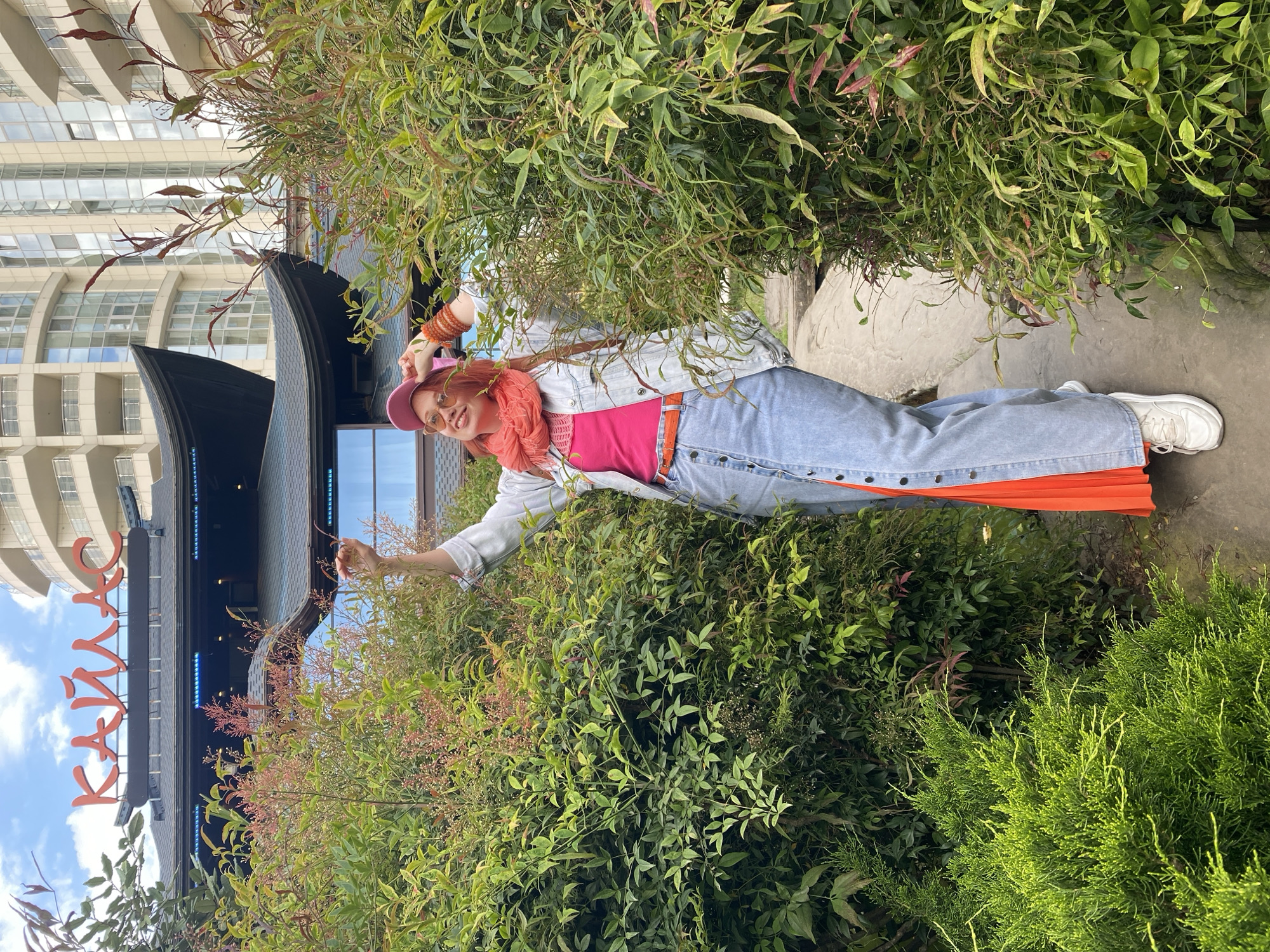 A woman stands in a plant-filled area next to a building in Sochi, Russia. She is wearing a pink hat, orange scarf, pink shirt, light blue denim jacket, blue jeans, and white sneakers. Her hair is red. The building has a sign in Cyrillic that reads "Кандалакша". The sky is visible in the upper left corner of the frame. The area is overgrown with various types of plants and bushes.
